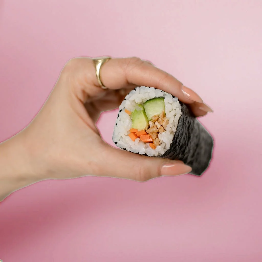 A hand holding a sushi roll with cucumber, carrots, tuna, and rice against a pink background.