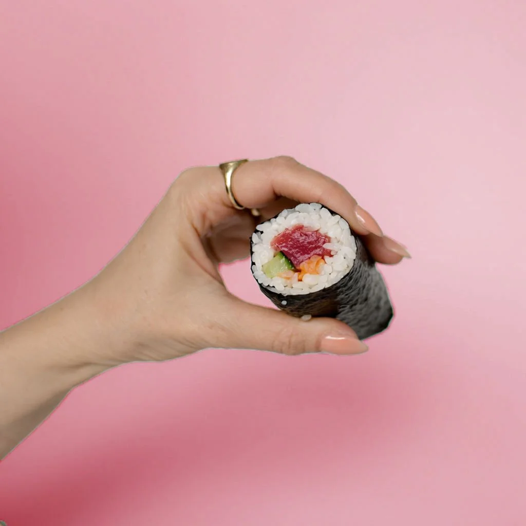 Person holding a sushi roll with tuna, avocado, and rice on a pink background.