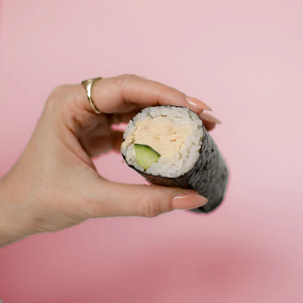 A hand holding a sushi roll with visible rice, cucumber, and yellow filling against a pink background.