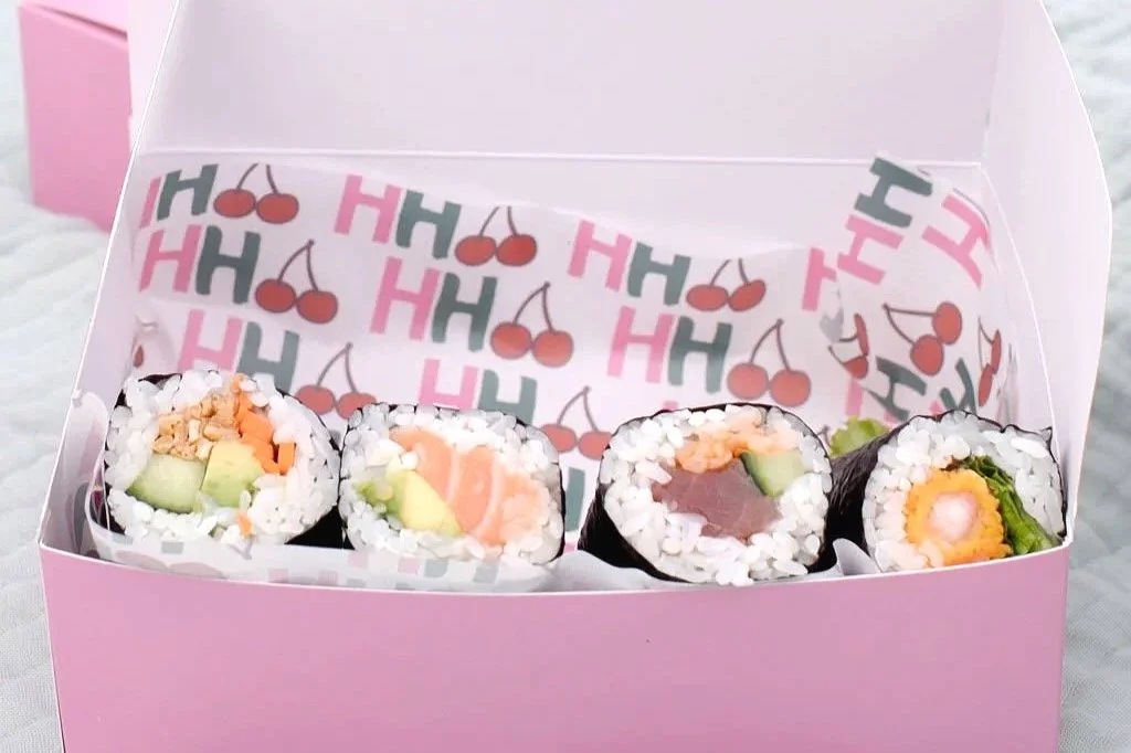 Assorted sushi rolls in a pink box with cherry and letter pattern liner inside.