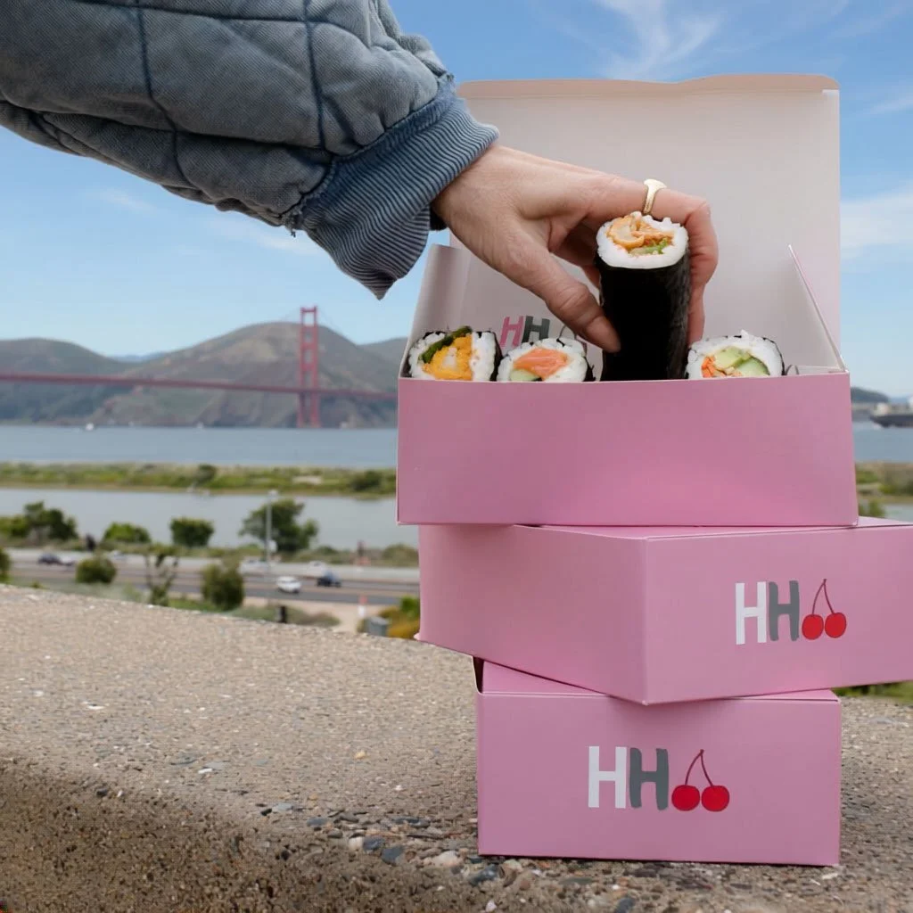 A person holding a sushi roll above pink boxes with cherries and the letters HH on them, with the Golden Gate Bridge and a body of water in the background.