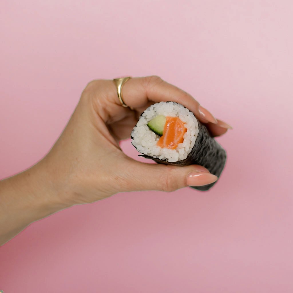 Close-up of a hand holding a sushi roll with cucumber and salmon inside, against a pink background.