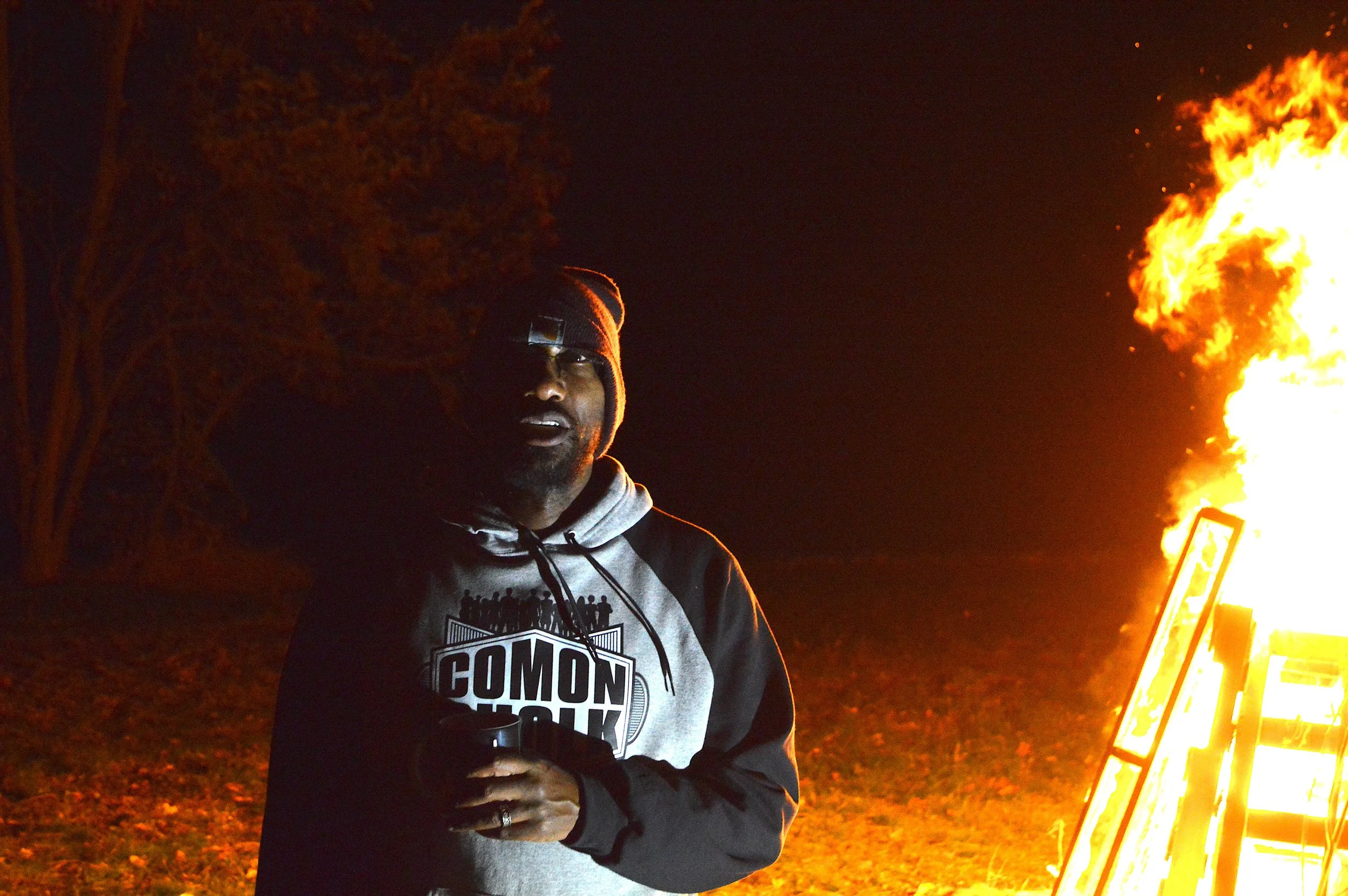 Person wearing a hoodie and glasses standing near a large fire at night in an outdoor area with trees, holding a cell phone.