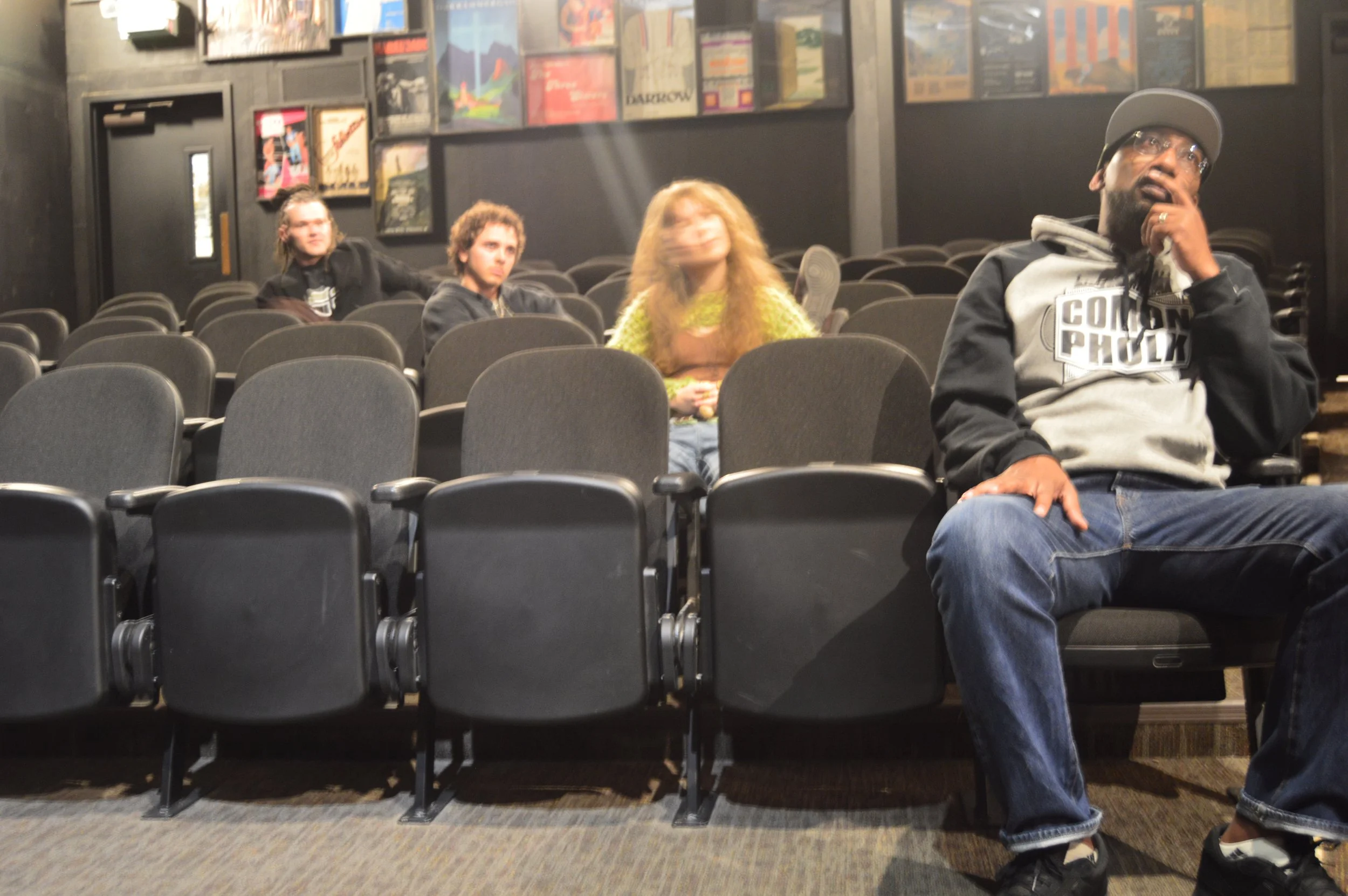 Four people sitting in an auditorium with black chairs and a dark wall covered with movie posters. One man in the foreground wears a gray hoodie, a baseball cap, and glasses. Two women and one man sit farther back, one woman has curly hair and a yellow top, the other woman has straight hair and a dark top, and the man has long hair. The image is slightly blurry.