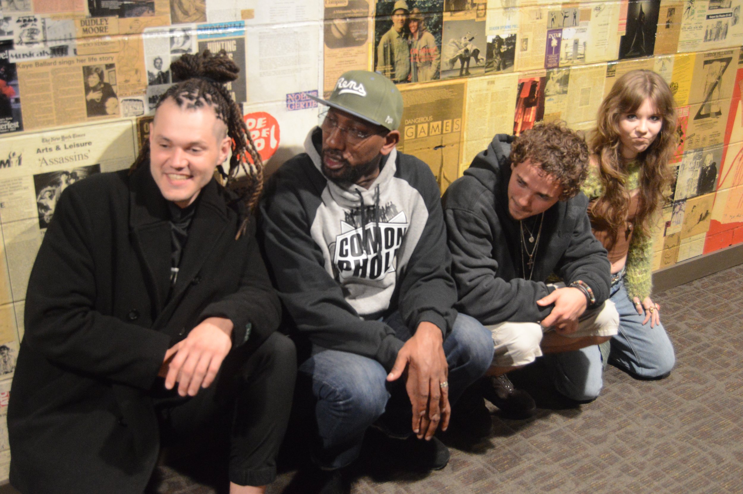 A group of five young people sitting on the floor against a wall decorated with vintage posters and newspaper clippings, some smiling and making playful faces.