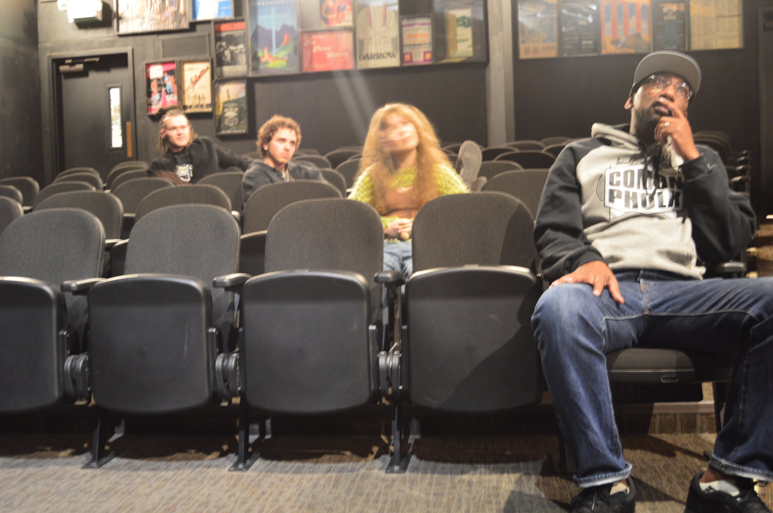 People seated in a dark theater with rows of black chairs, some looking towards the camera and others looking away, with wall art and posters in the background.