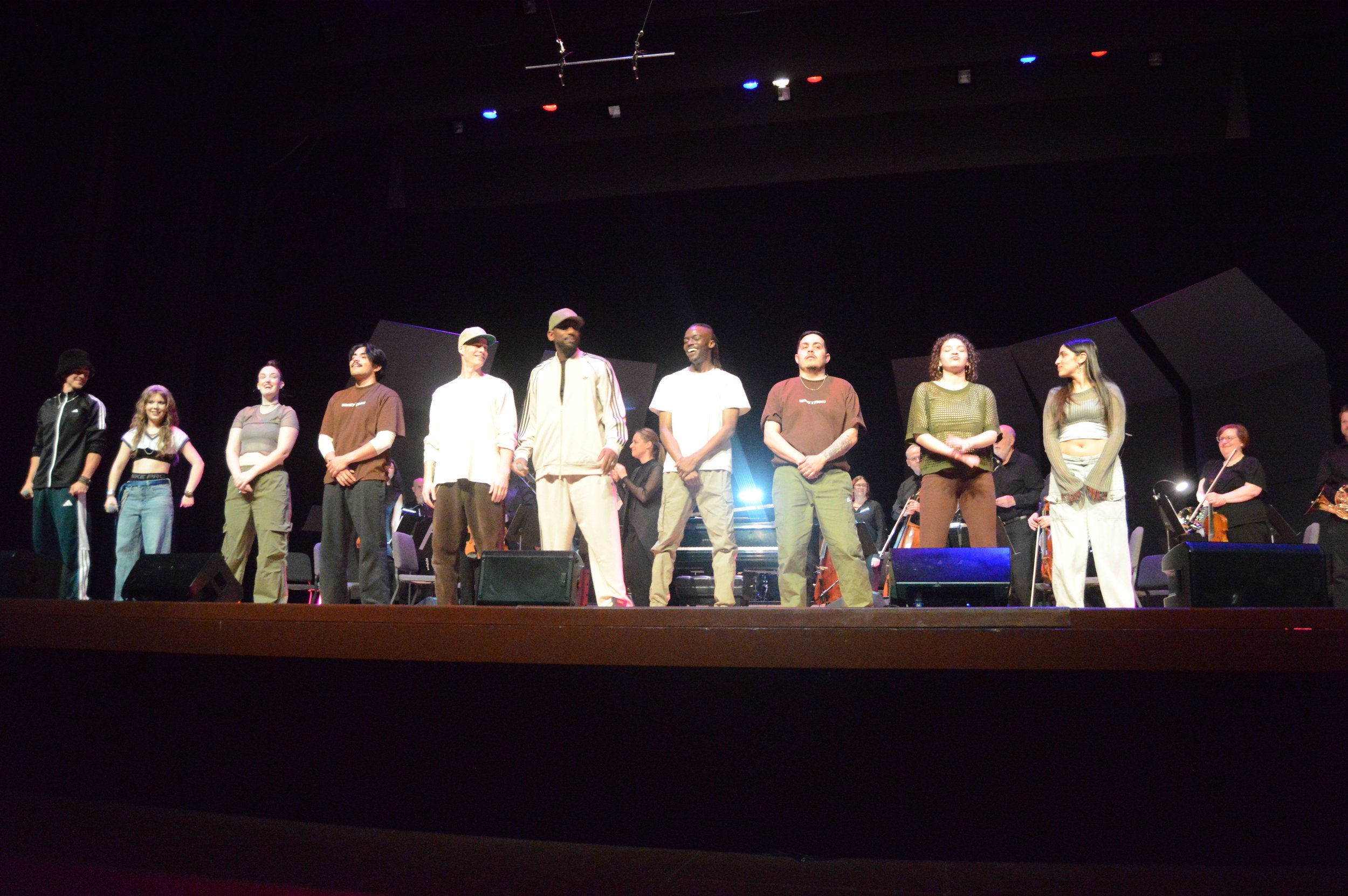 A diverse group of performers and musicians standing on a stage during a concert or rehearsal, with some holding microphones and others with musical instruments, under stage lighting.