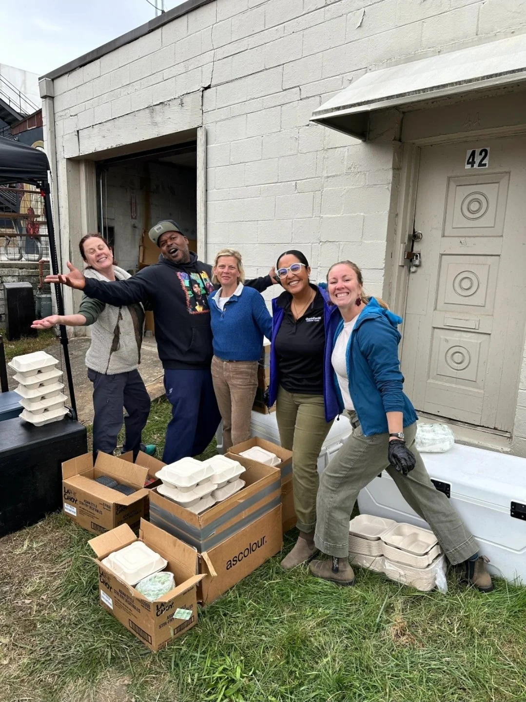 Group of five people smiling including the E1T1 team and posing outdoors, with boxes of food containers in front of them, standing next to a building with a white brick wall, preparing to deliver them to families immediately after Hurricane Helene.