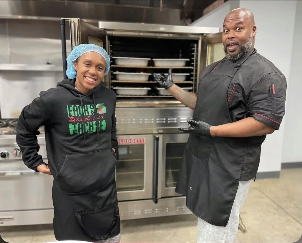A young woman and a man representing E1T1 wearing black aprons and gloves, standing in front of an industrial oven in a commercial kitchen. The woman is smiling, and the man is pointing towards the oven with a surprised expression.