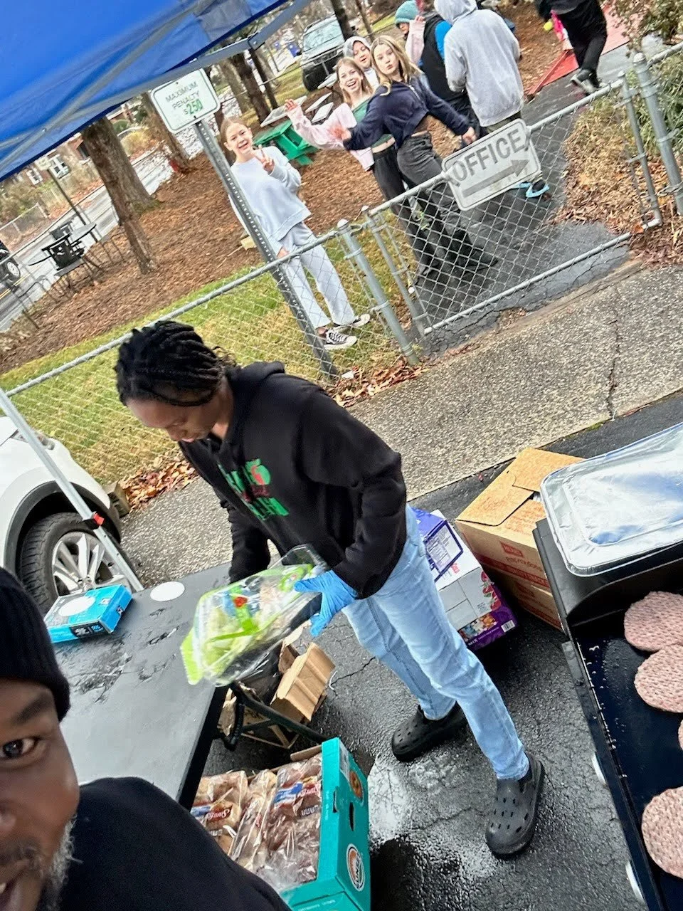 A person in black hoodie preparing food at an outdoor event with people standing behind a fence, and a sign that says 'Office' on the fence.