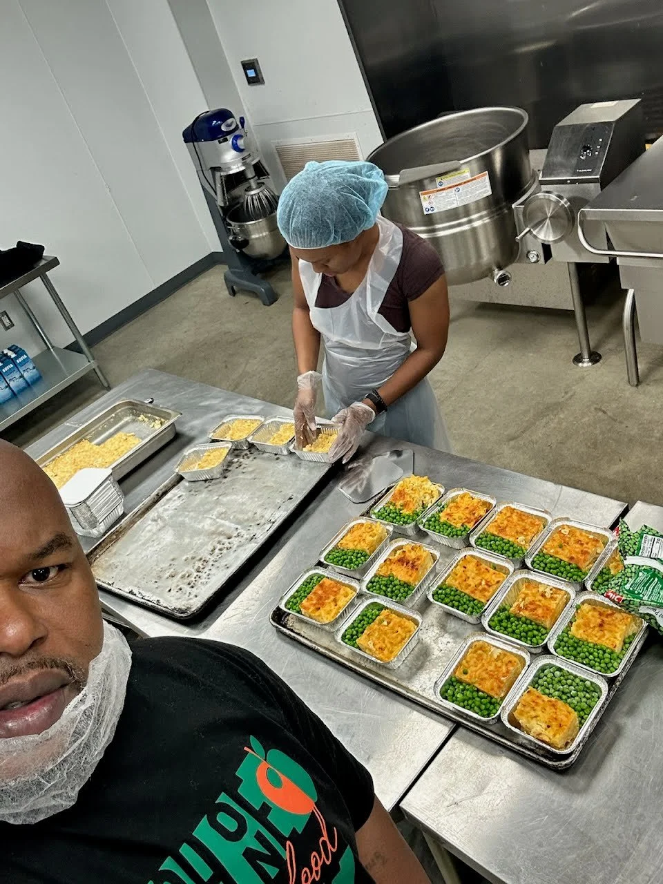 Two people preparing and packing trays of food in a commercial kitchen. One person is taking a photo, and the other person is arranging food in aluminum trays that contain a casserole dish with green peas on the side.