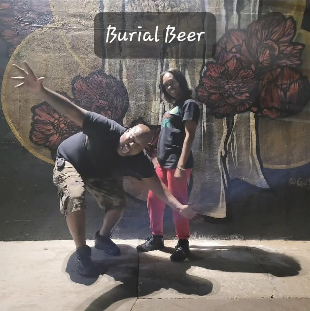 E1T1 Co-Founders posing in front of a mural paint art of large flowers with the text 'Burial Beer' on it. The person on the left is bending forward and pointing, while the person on the right is standing and smiling.
