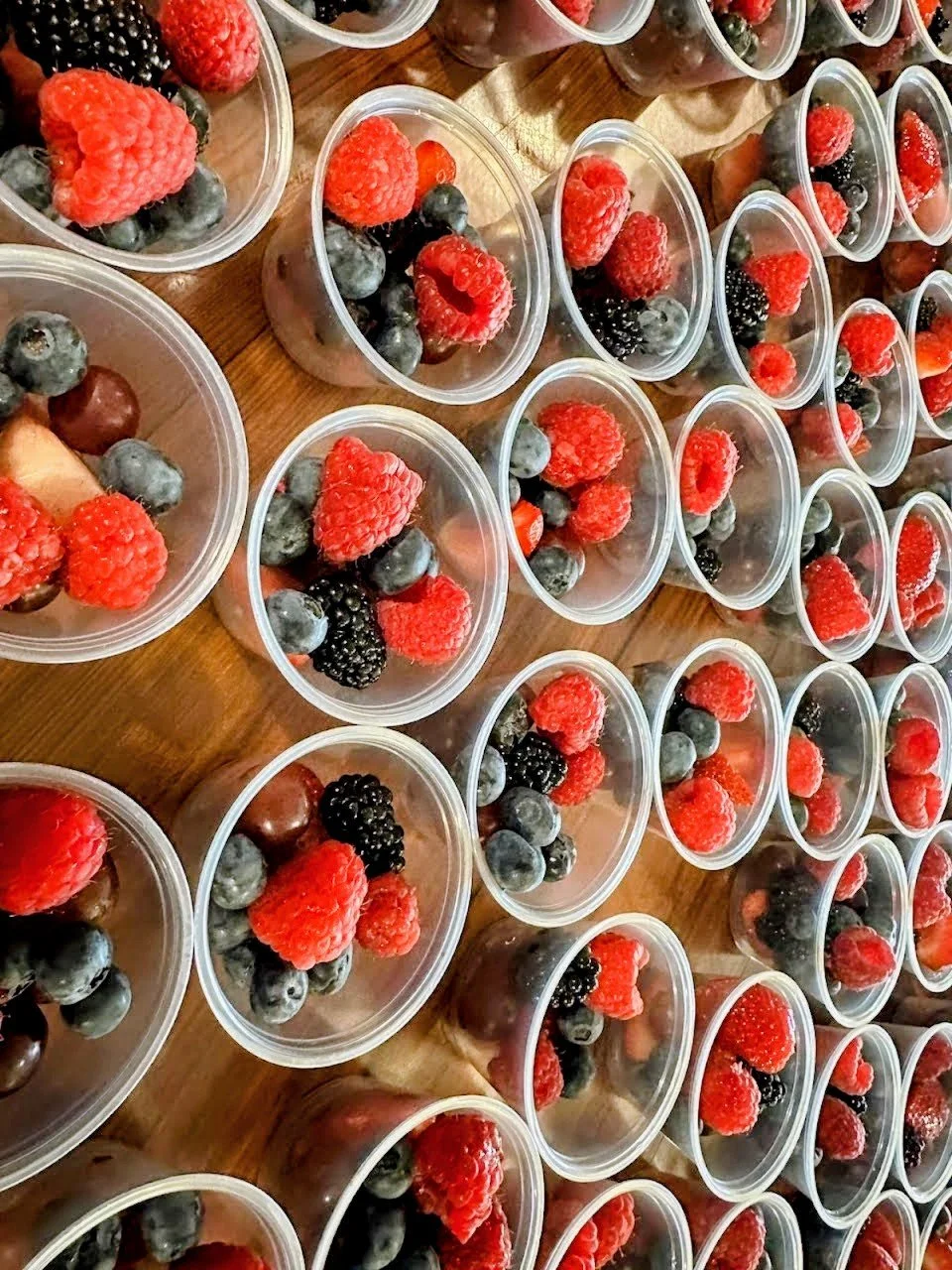 Rows of clear plastic cups filled with mixed berries, including fresh raspberries, blueberries, blackberries, and strawberries, placed on a wooden surface.