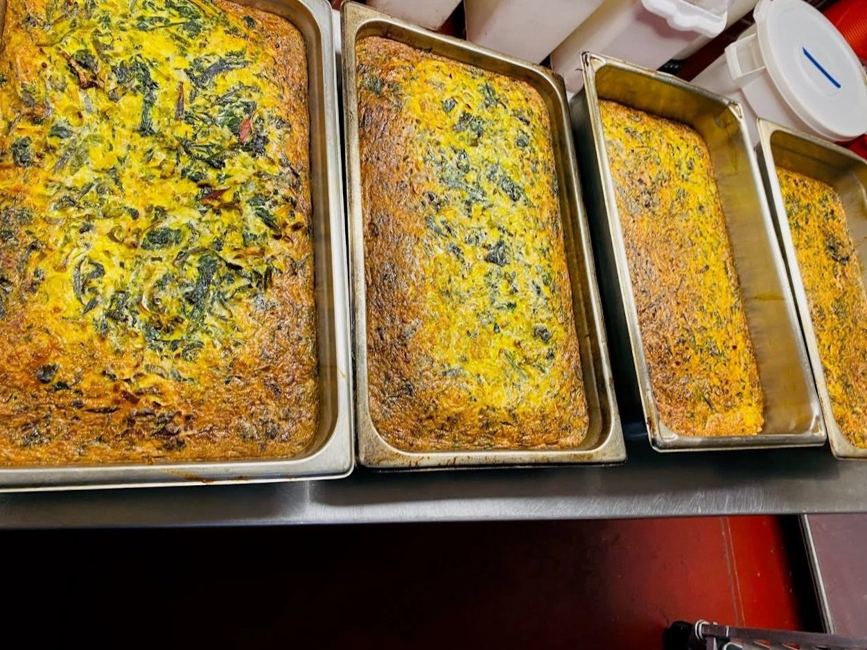 Four trays of baked casserole dishes with layers of vegetables and herbs, topped with melted cheese, in a commercial kitchen setting.