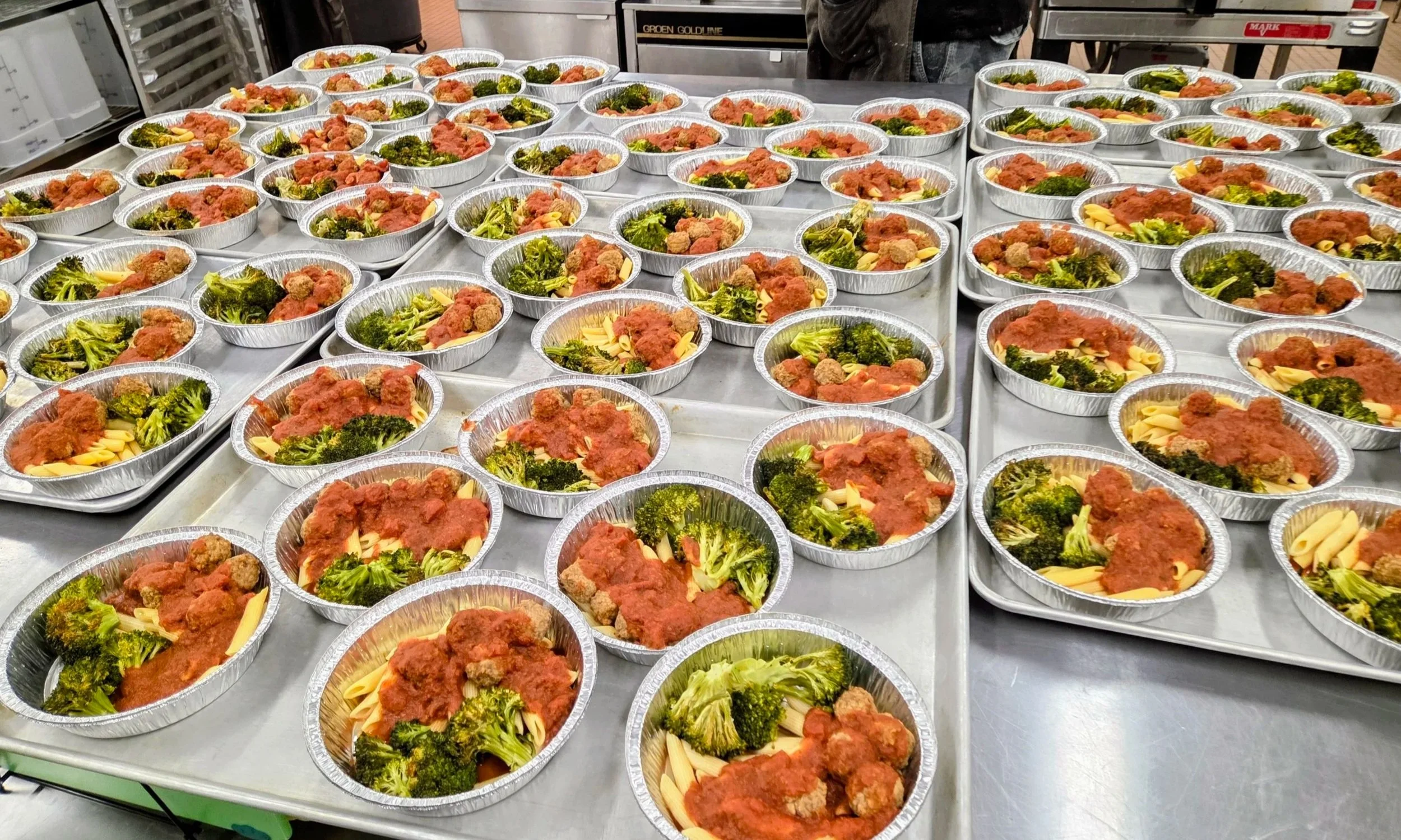 Multiple aluminum trays of cooked pasta, broccoli, and meatballs with tomato sauce on a stainless steel table at a food service setting.