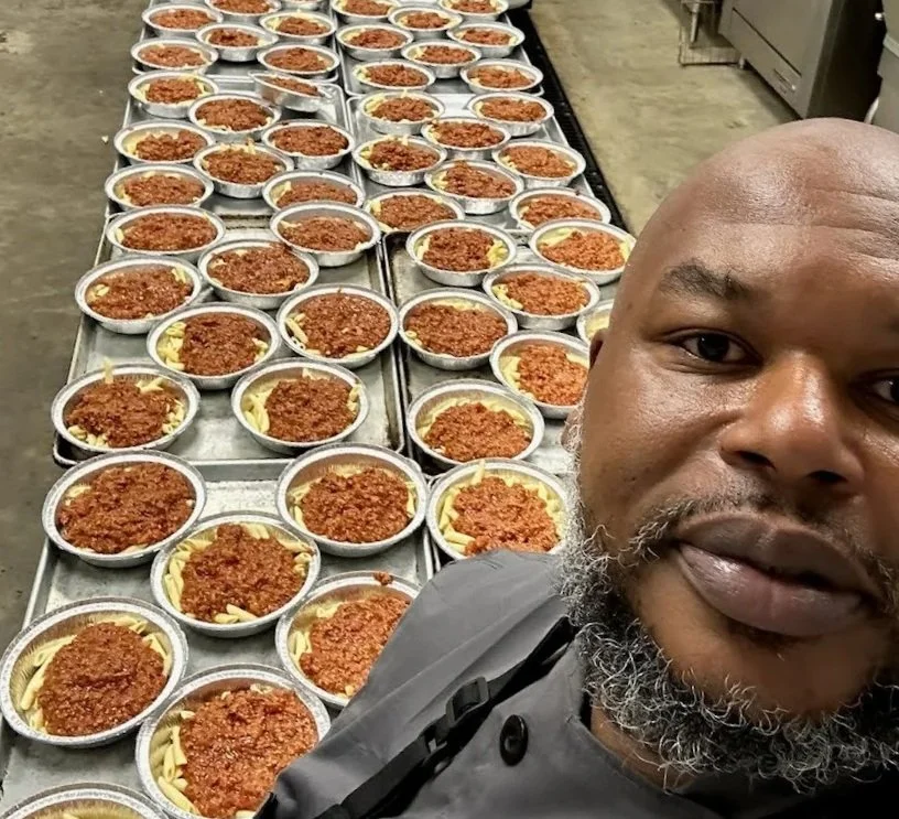 E1t1 Chef takes a selfie in a commercial kitchen with numerous bowls of spaghetti and meat sauce prepared and laid out on a stainless steel table.