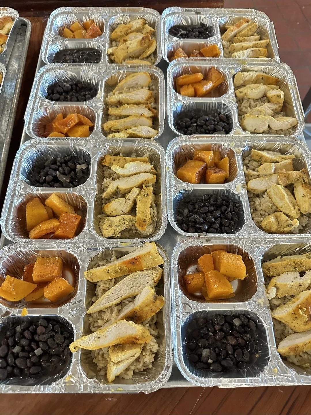 Multiple aluminum foil meal containers with black beans, cooked carrots, and sliced cooked chicken on a wooden table.