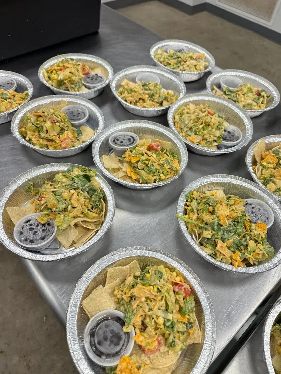 Multiple aluminum foil takeaway containers filled with chopped vegetable salads and tortilla chips, placed on a metal table.