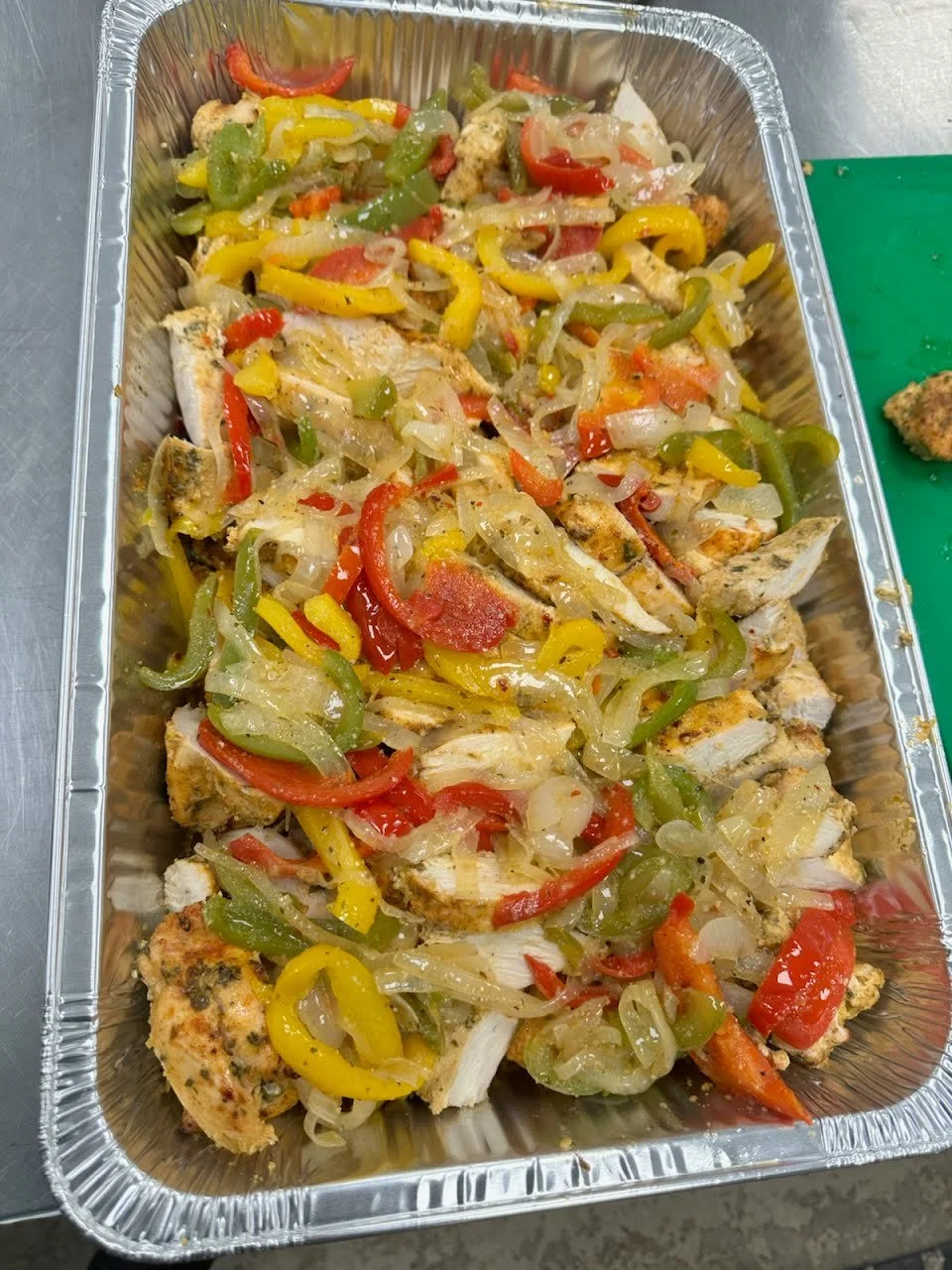 Cooked chicken with sautéed red, yellow, and green bell peppers and onions in a foil tray.