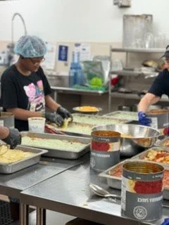 People preparing food in a commercial kitchen, wearing hair nets and gloves, with various ingredients and containers on the counter.