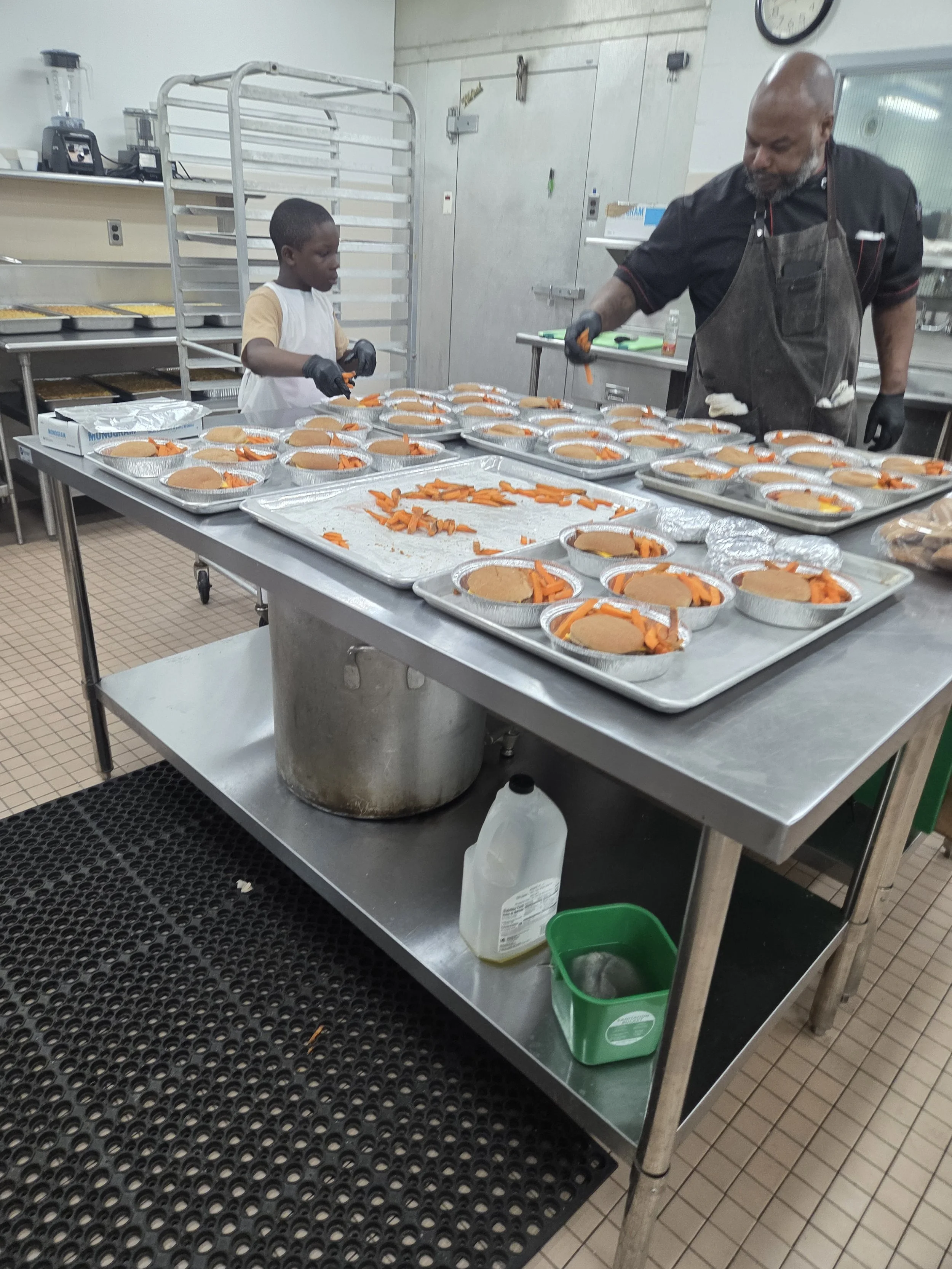 A kitchen or food preparation area with a stainless steel table holding trays of sliced carrots and baked goods. A man and a boy are working together, preparing food, both wearing black gloves. The boy is slicing food, and the man is holding a knife. There are trays with food in the background, along with kitchen appliances, a clock, and various kitchen utensils.