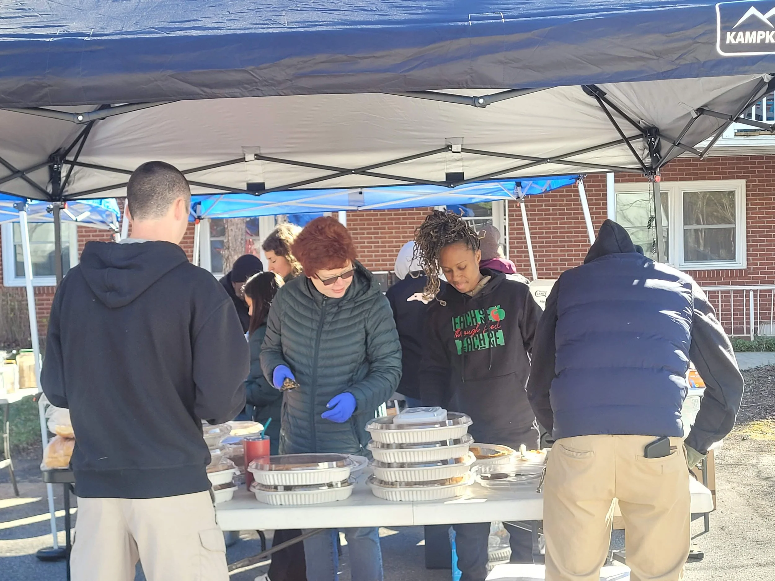 E1T1 and their volunteers and supporter gathered under a canopy, serving food at an outdoor community event on a sunny day.