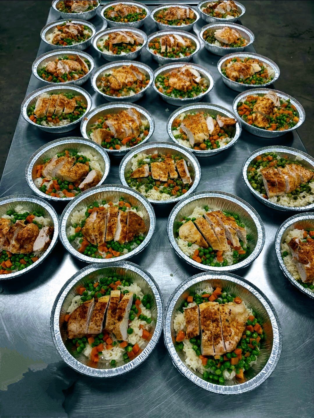 Multiple aluminum containers filled with cooked chicken breast slices, seasoned rice, and mixed vegetables including carrots and peas, arranged on a stainless steel surface.