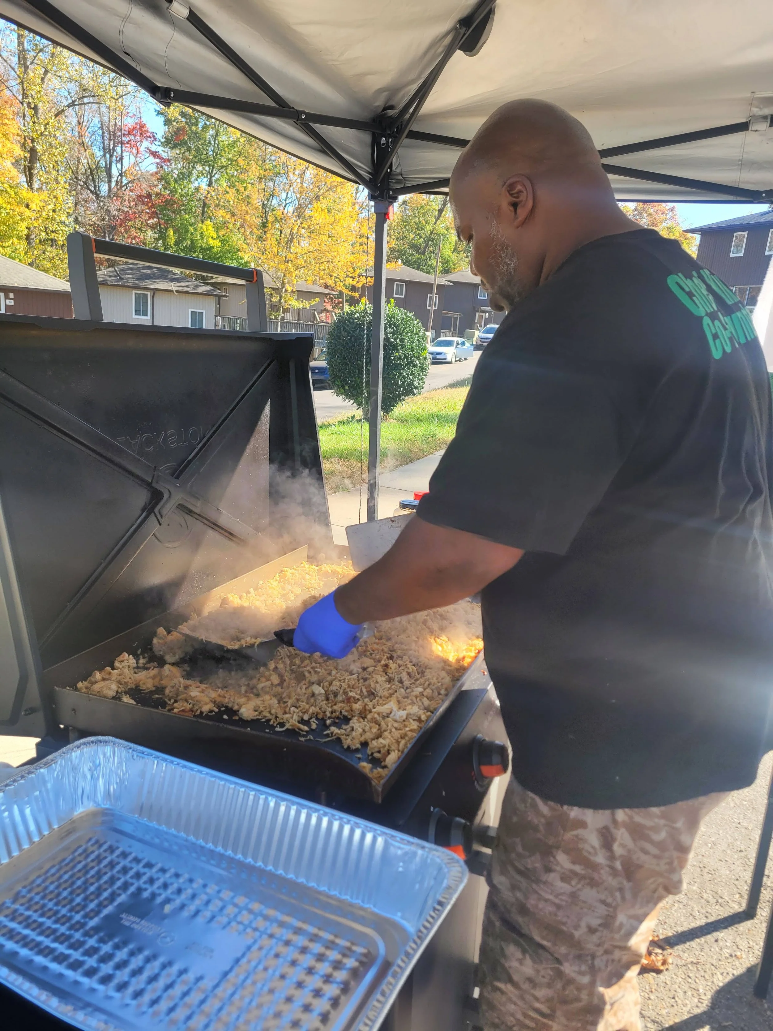 An E1T1 Chef wearing a black T-shirt and camouflage shorts is preparing food on a grill outdoors under a canopy, with trees and houses in the background in neighborhood during the aftermath of Hurricane Helene.