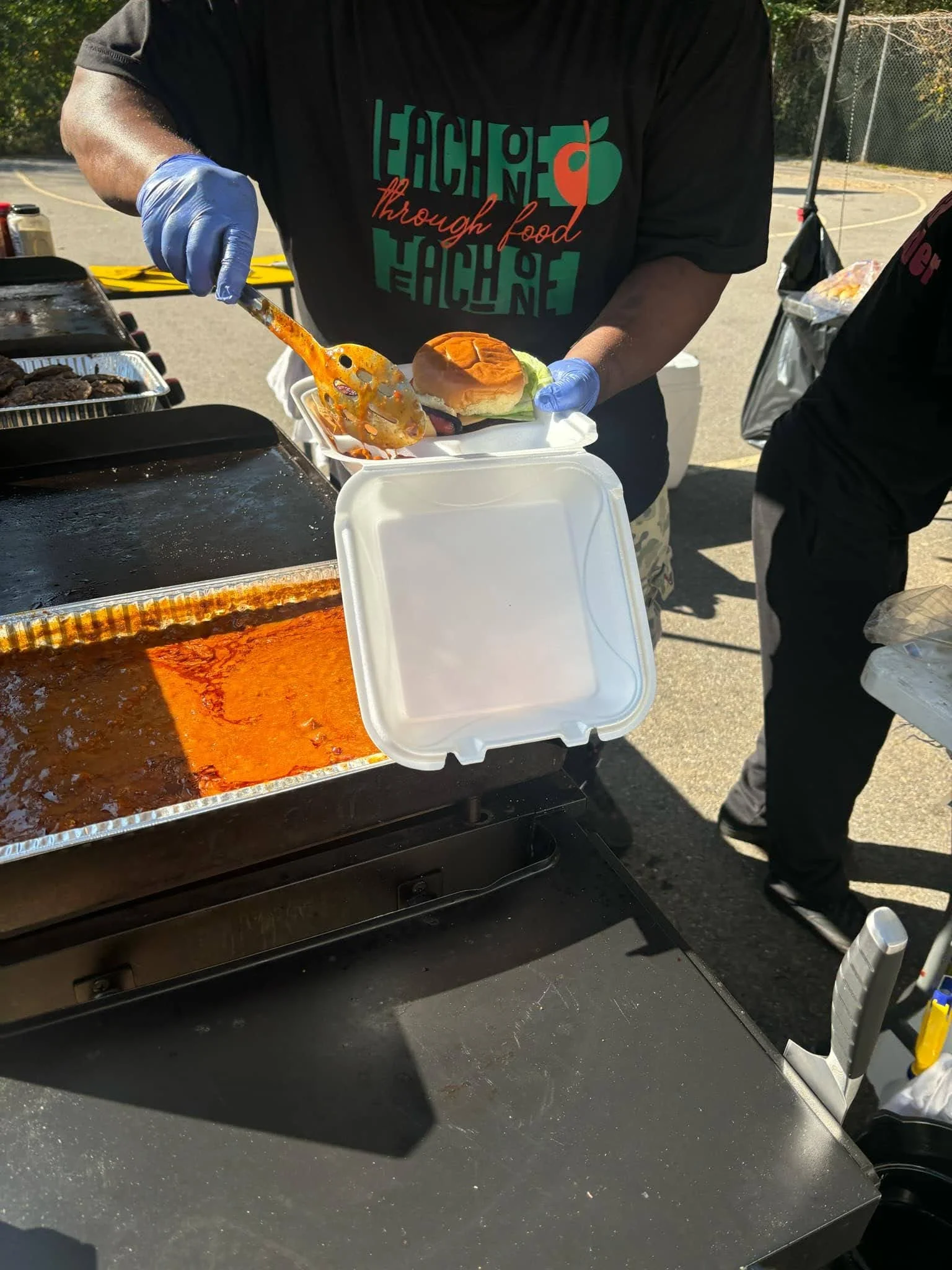 E1T1 Chef wearing a black t-shirt with colorful text and a graphic holding a burger and an orange spatula, serving food at an outdoor event. There is a tray of chili, and a table with condiments, in a neighborhood during the aftermath of Helene.