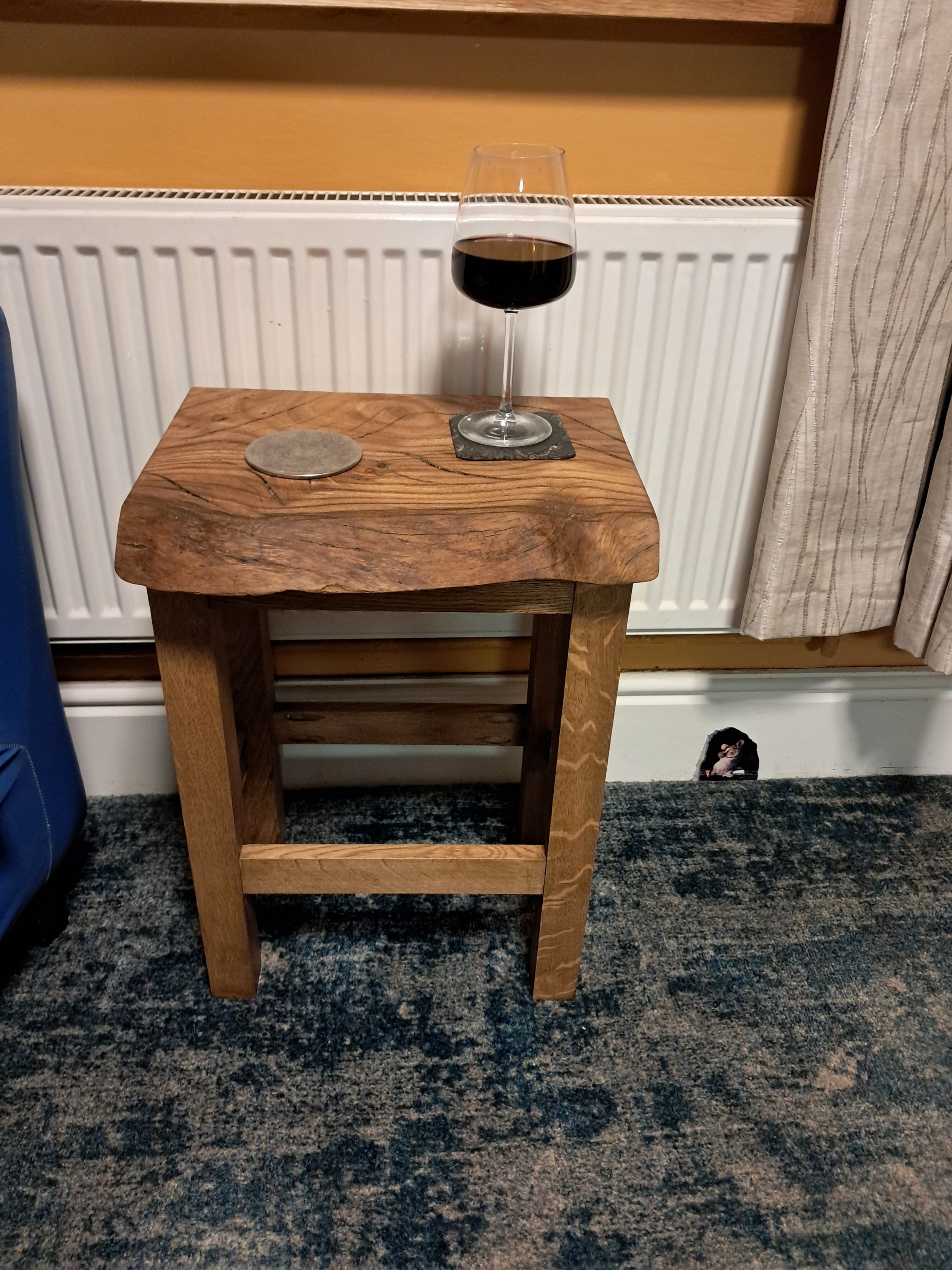 A small wooden table with a glass of red wine on a black coaster, placed in front of a radiator. There is a gray stone coaster beside the glass and a small piece of paper or sticker on the carpet below the table.
