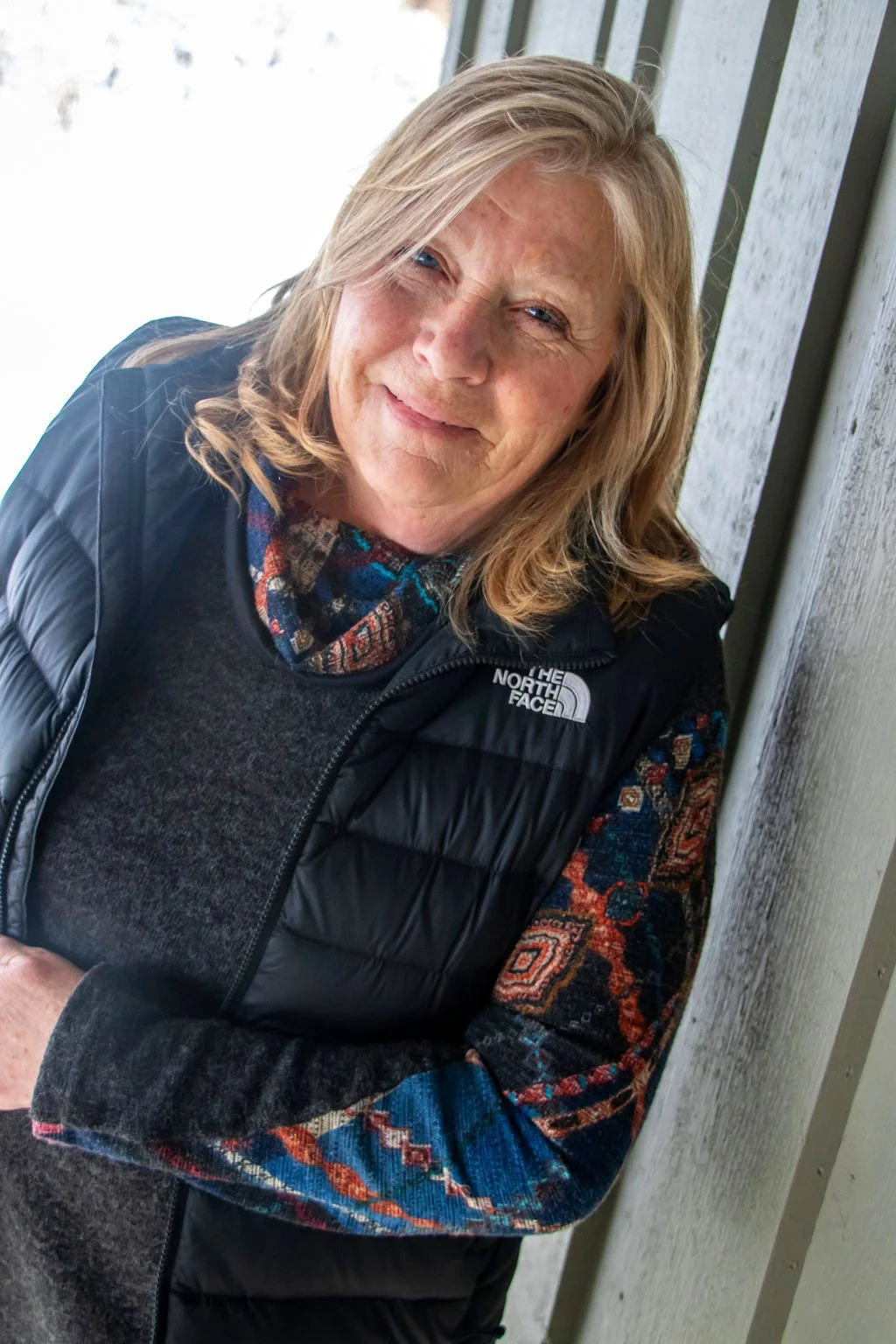 A smiling woman with blonde hair and blue eyes, wearing a patterned shirt, dark sweater, and a black North Face vest, sitting outdoors near a wooden wall.