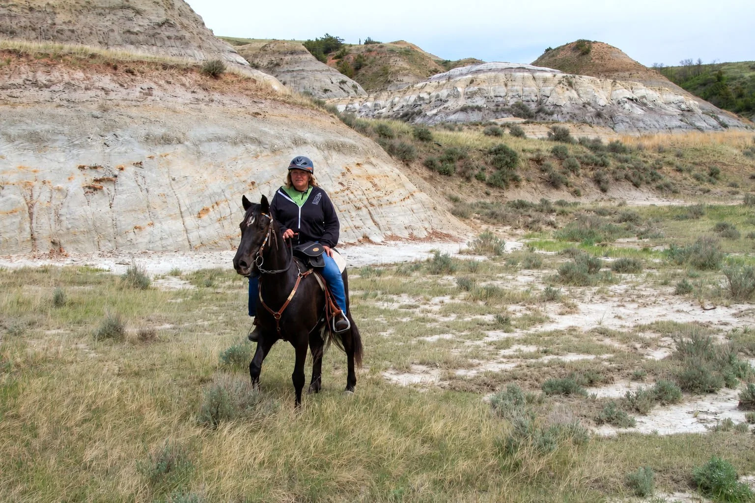 A woman riding a black horse in a desert landscape with eroded white and beige cliffs and sparse green vegetation.