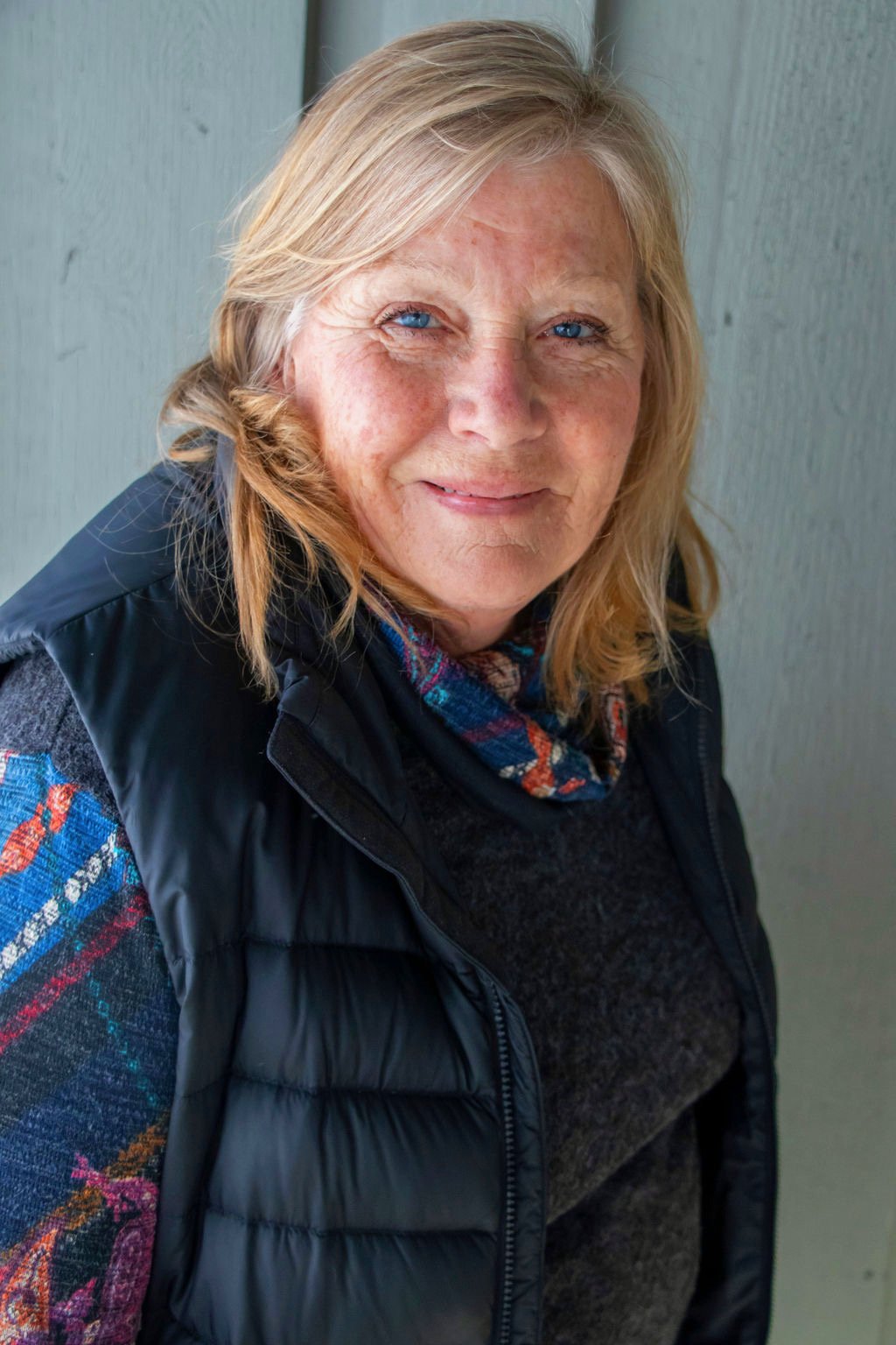 A woman with blonde hair wearing a plaid scarf, a dark gray sweater, and a black puffy vest, standing against a light-colored background.
