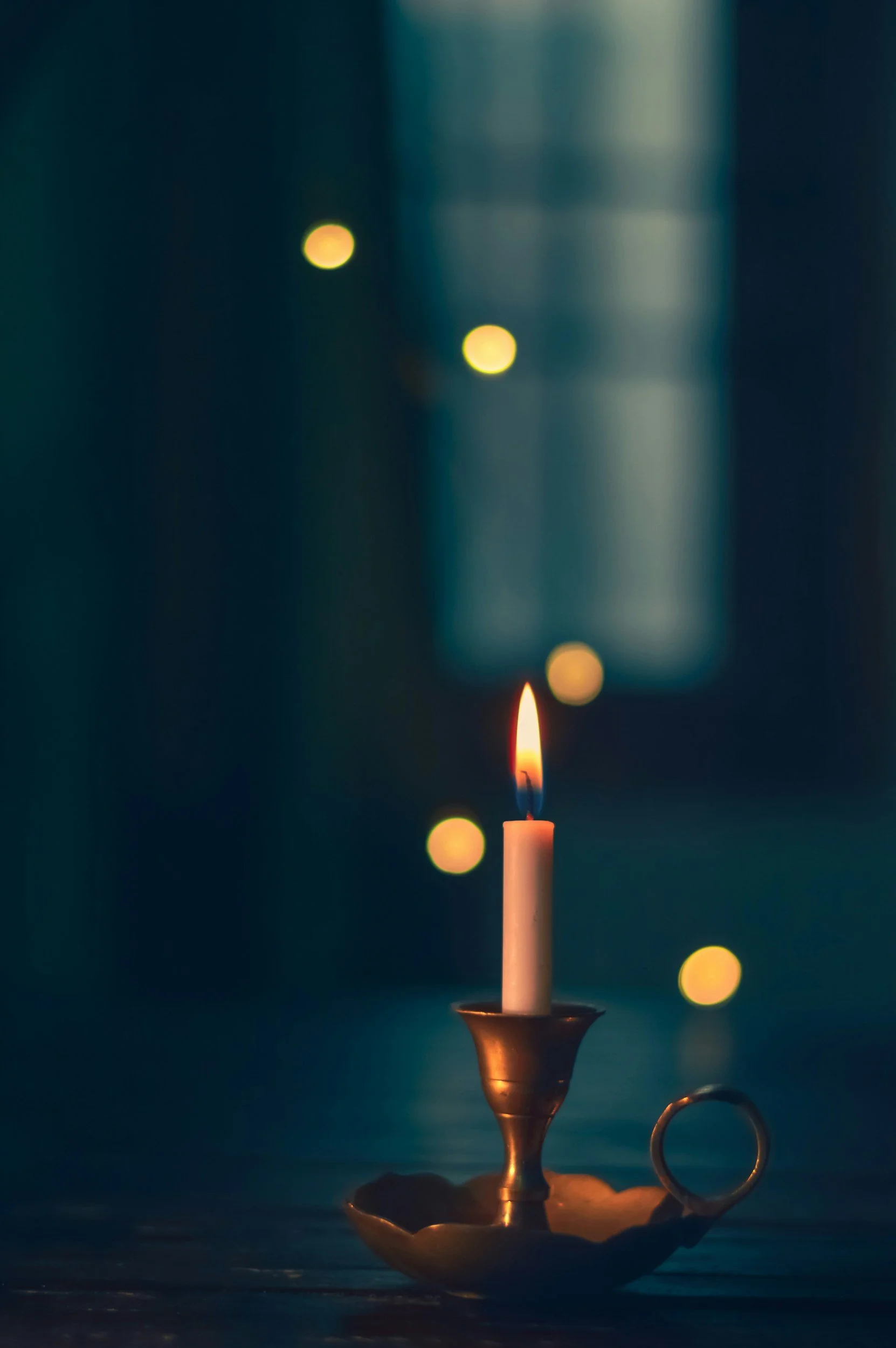 A lit candle in a brass holder with a curved handle, placed on a dark surface. The background is dark with blurred yellow and white bokeh lights, creating a warm, calm atmosphere.