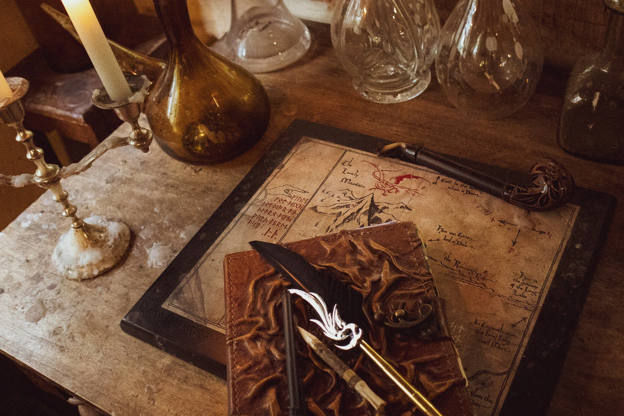 A desk with a vintage map, a black and gold ballpoint pen, glass vases, and a candle holder with a single candle. The map appears to be an illustrated map of the Lord of the Rings.
