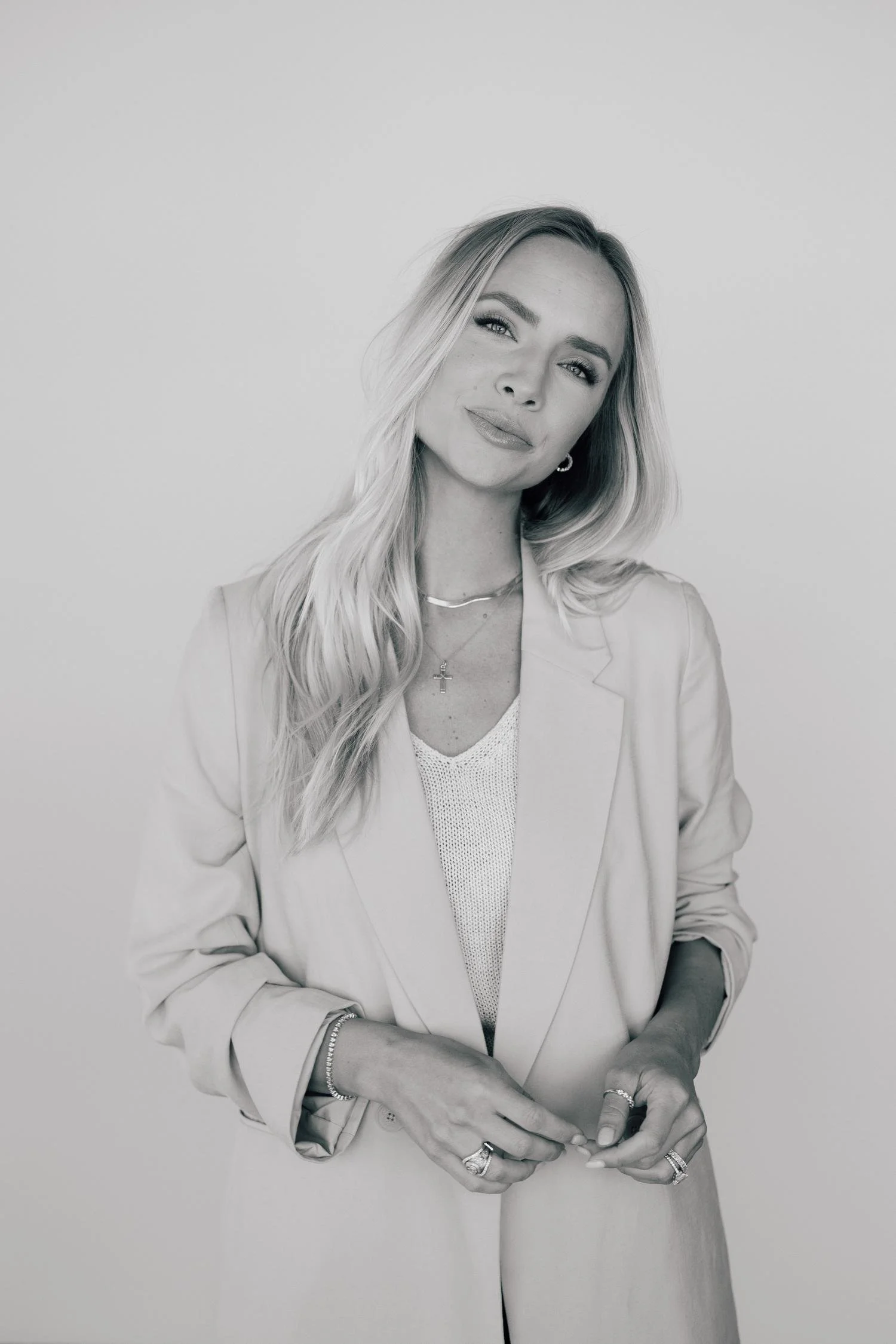 Black and white portrait of a woman with long, wavy hair, wearing a blazer, jewelry, and a confident smile, standing against a plain background.
