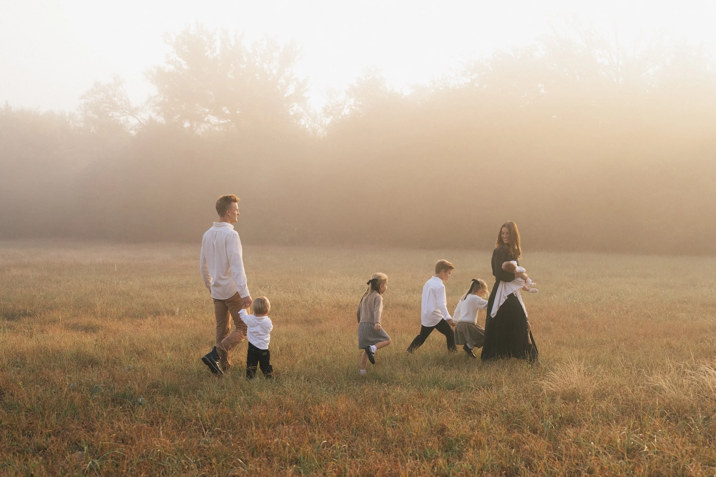 A family walking through a grassy field during sunset, with trees in the background and a woman holding a baby.