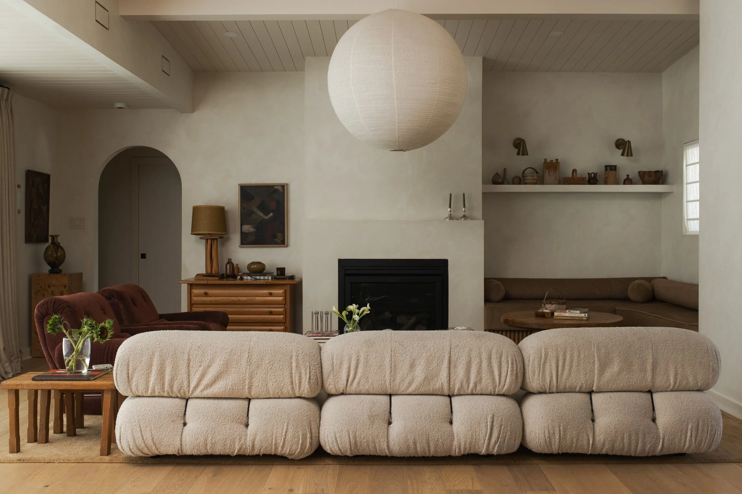 A cozy living room with a large, rounded off-white plush ottoman in the foreground, surrounded by a beige sofa and a burgundy armchair. There are wooden side tables, a lamp, artwork on the walls, a fireplace, and decorative objects on shelves, with n