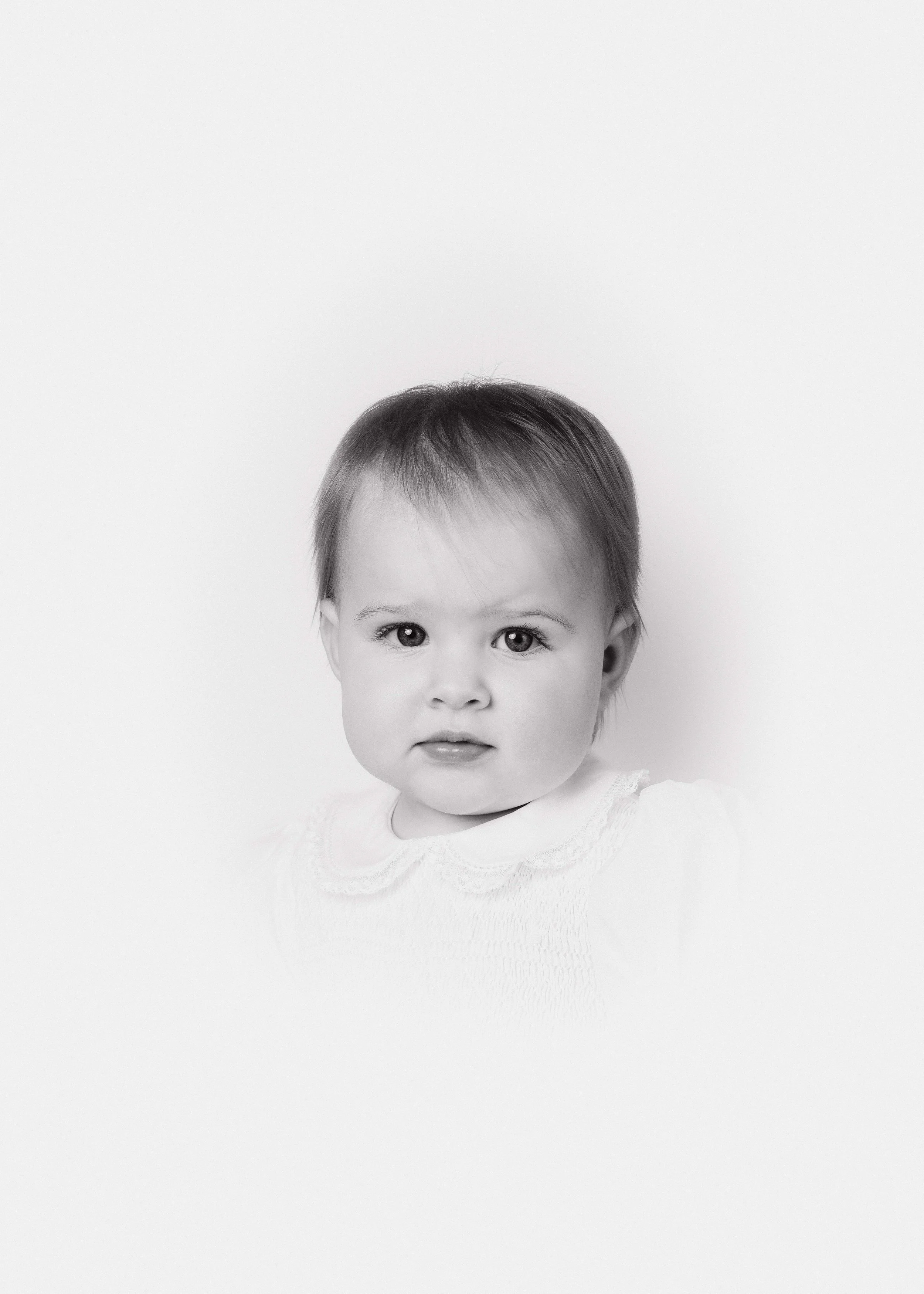 Black and white photo of a young child with short dark hair, wearing a light-colored top, looking directly at the camera with a neutral expression, against a plain white background.