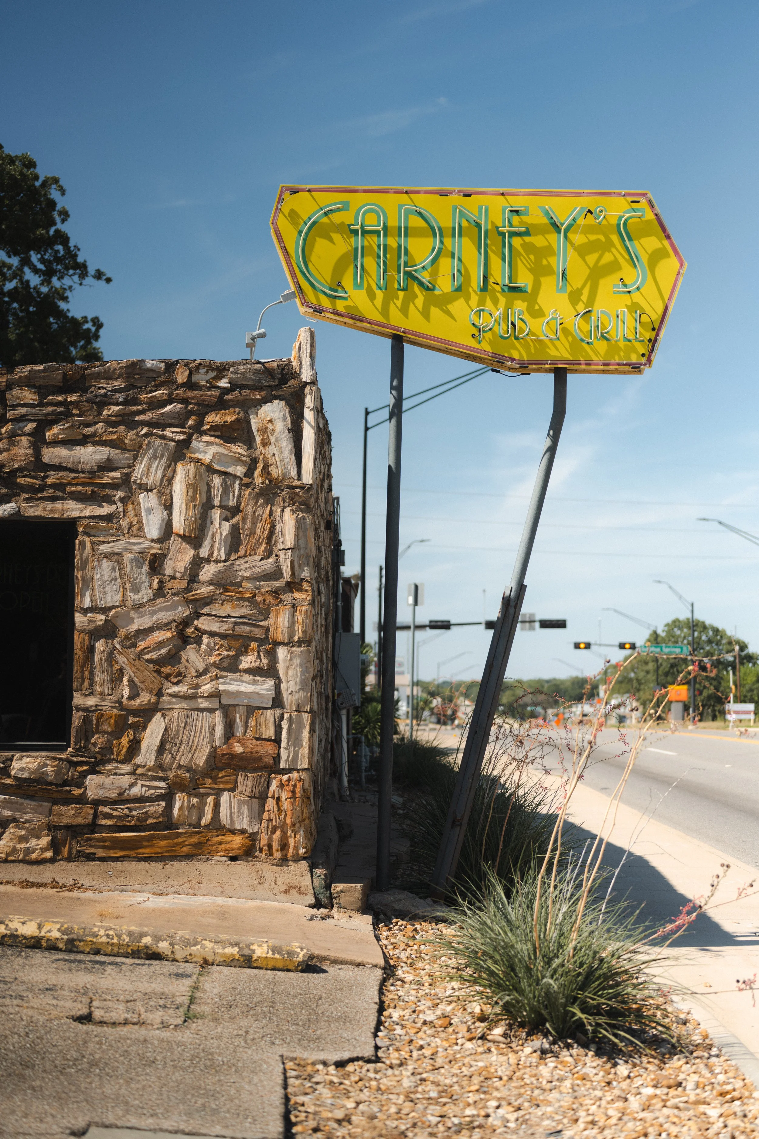 A yellow neon sign for 'Cirey's Pub & Grill' mounted on a metal pole outside a building with a stone facade. The sign is casting shadows and the sky is clear and blue. In the background, there are traffic signals and streetlights.