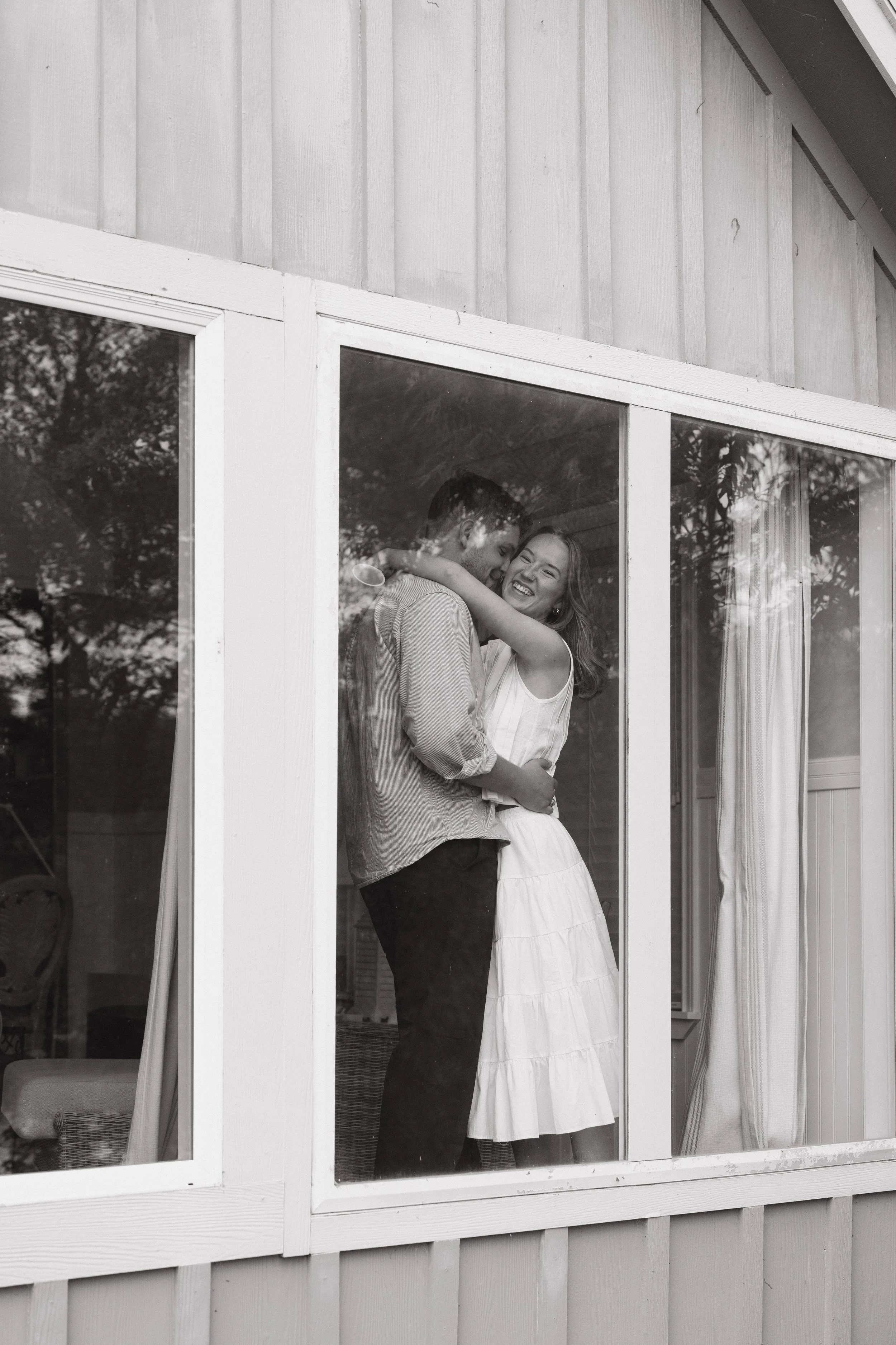 A black-and-white photo of a smiling woman in a white dress hugging a man in a striped shirt through a large glass window of a house with vertical siding.