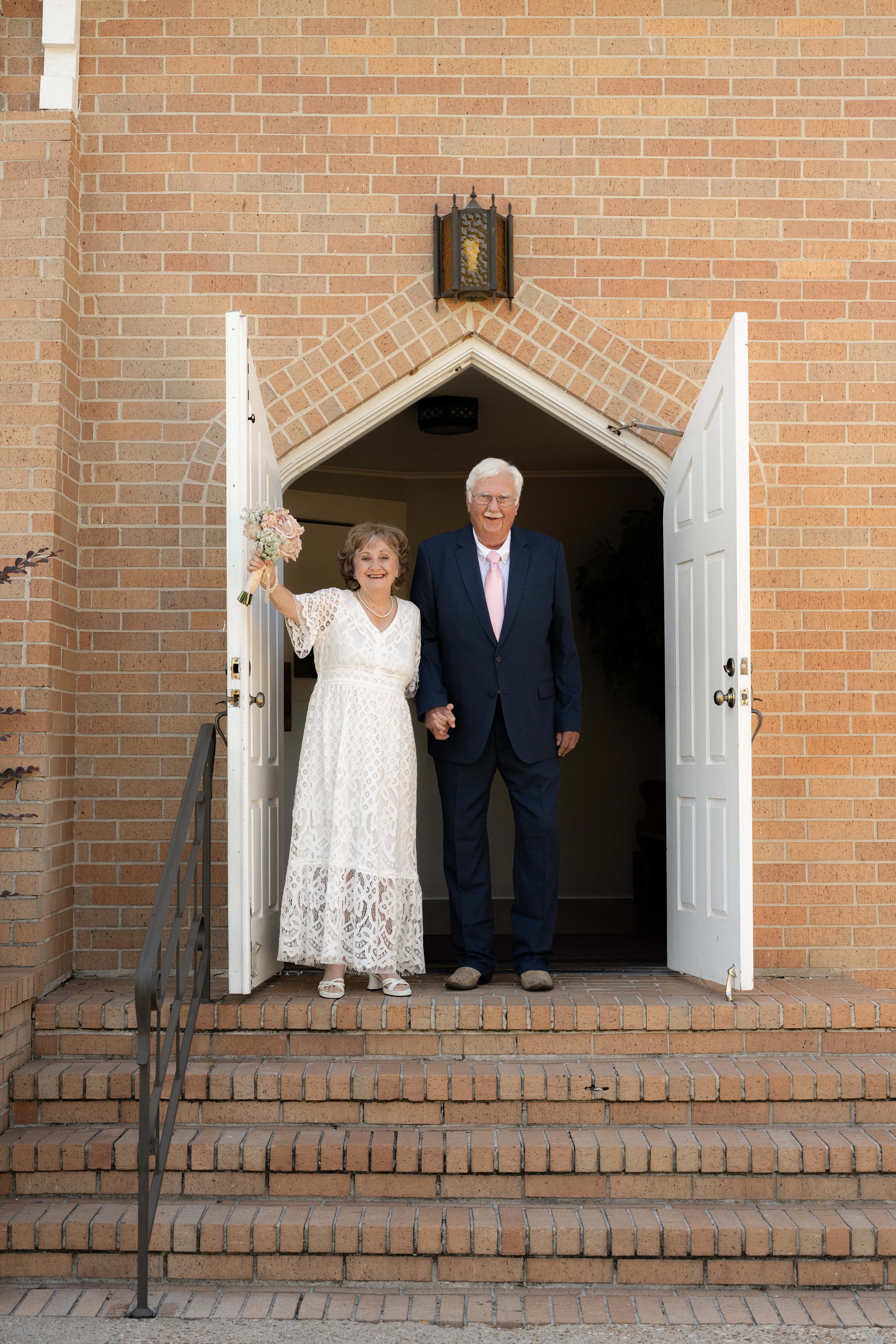 An elderly woman and man standing at the open doorway of a brick building, holding hands. The woman is wearing a white lace dress and the man is in a navy suit with a pink tie. The woman is holding a bouquet of flowers and smiling, while the man is also smiling. The scene appears to be a special occasion, possibly a wedding or celebration.
