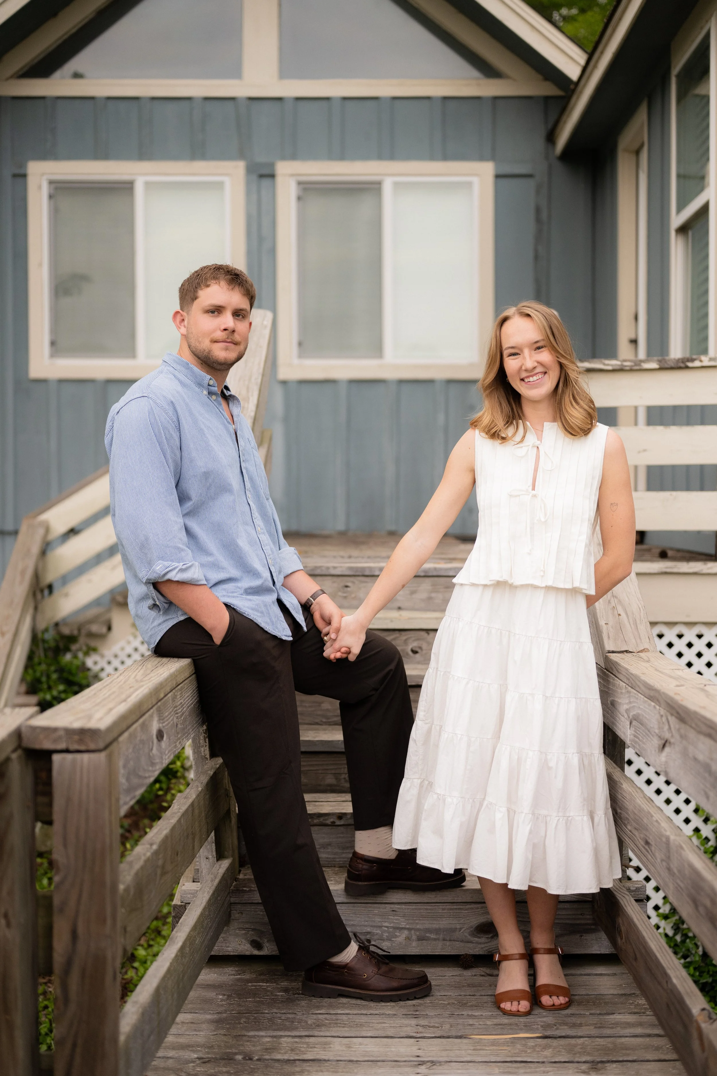 A young man and woman standing outdoors on a wooden staircase, holding hands, in front of a blue house with white window frames.