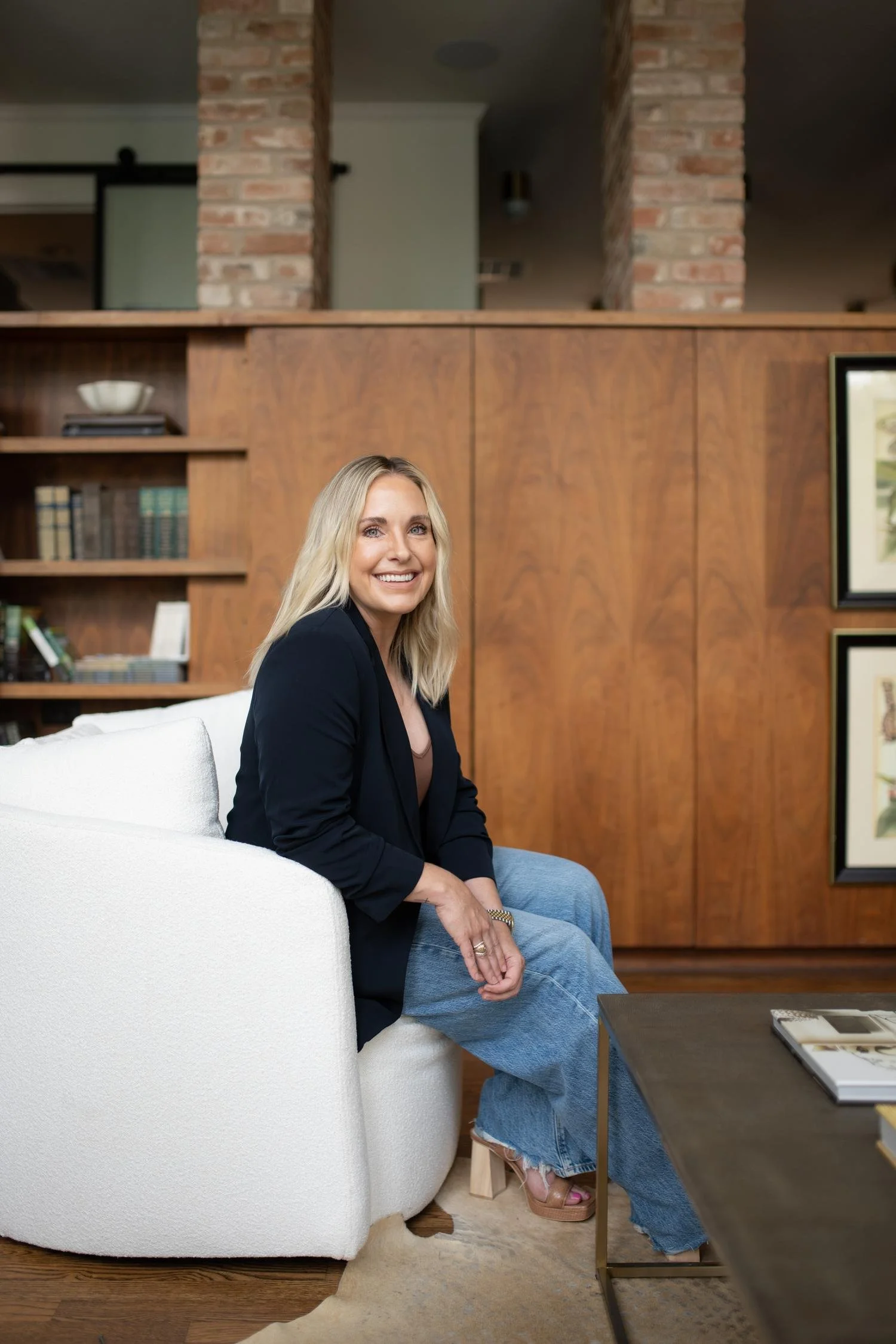 A woman with blonde hair smiling while sitting on a white sofa in a room with wooden panel walls and a brick column, with a coffee table in front of her.