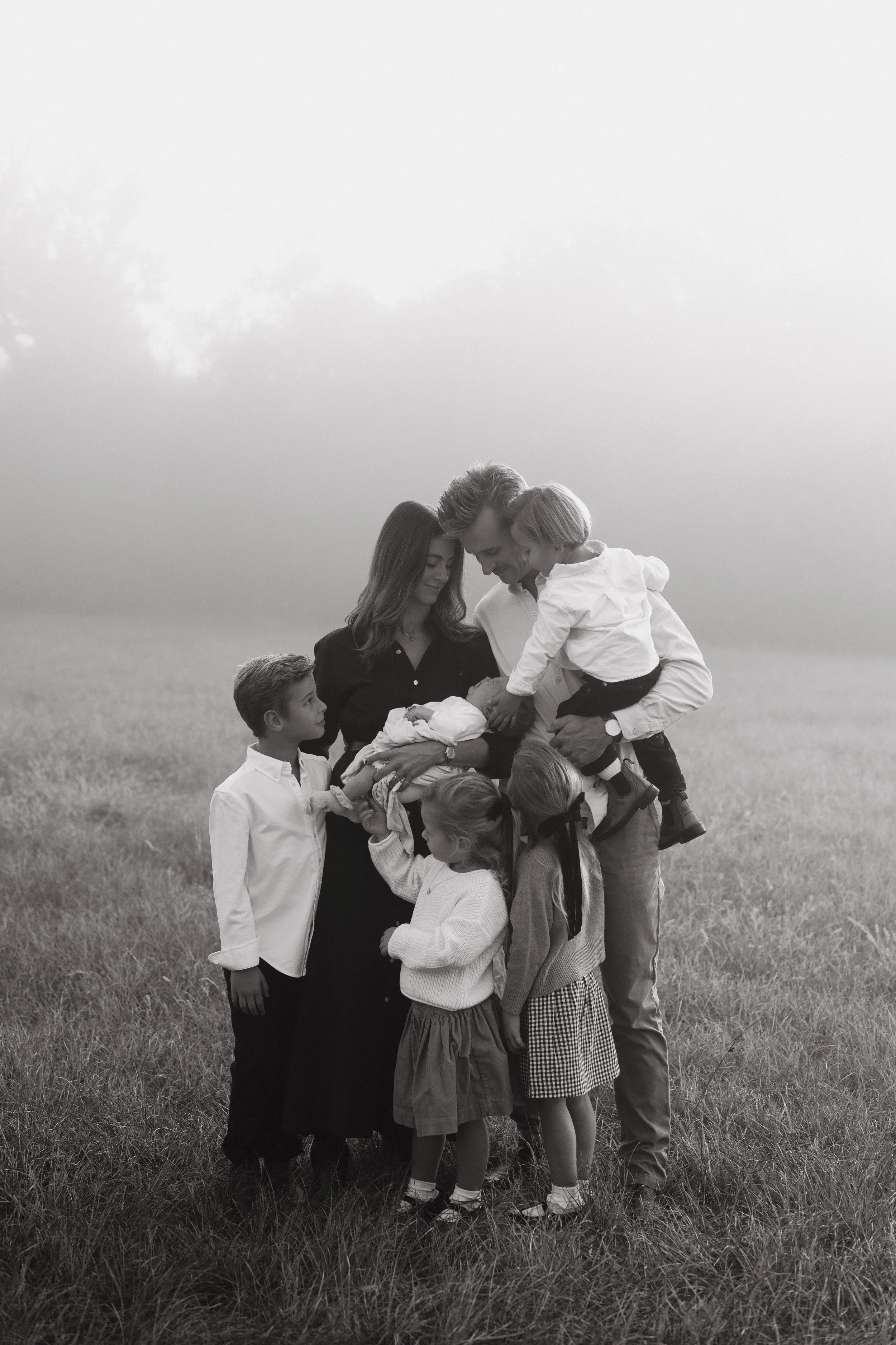 A black-and-white photo of a family outdoors in a grassy field, gathered around a newborn baby, with adults and children smiling and touching the baby.