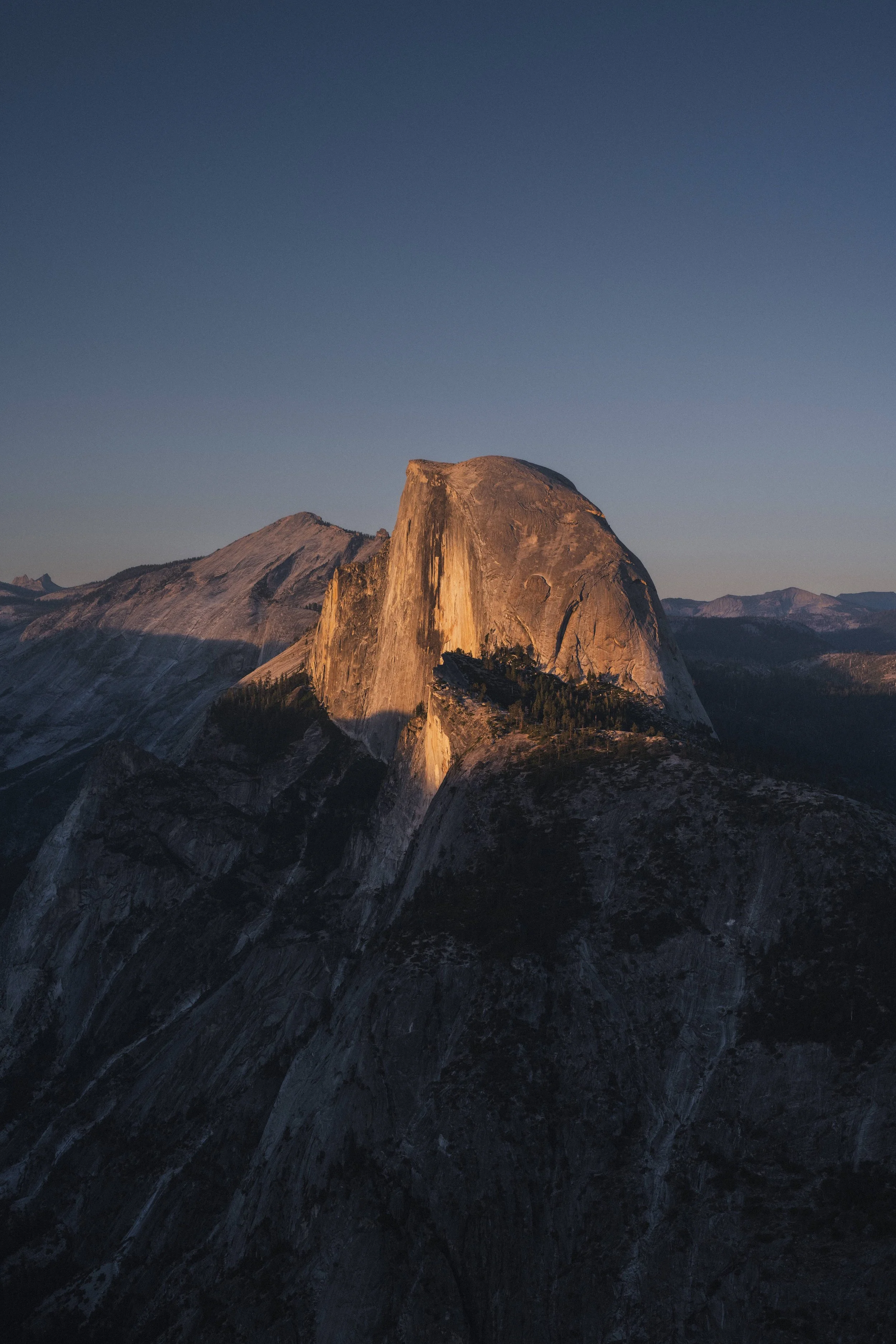 A large rocky mountain formation illuminated by the sunset, with a clear blue sky above and smaller mountains in the distance.