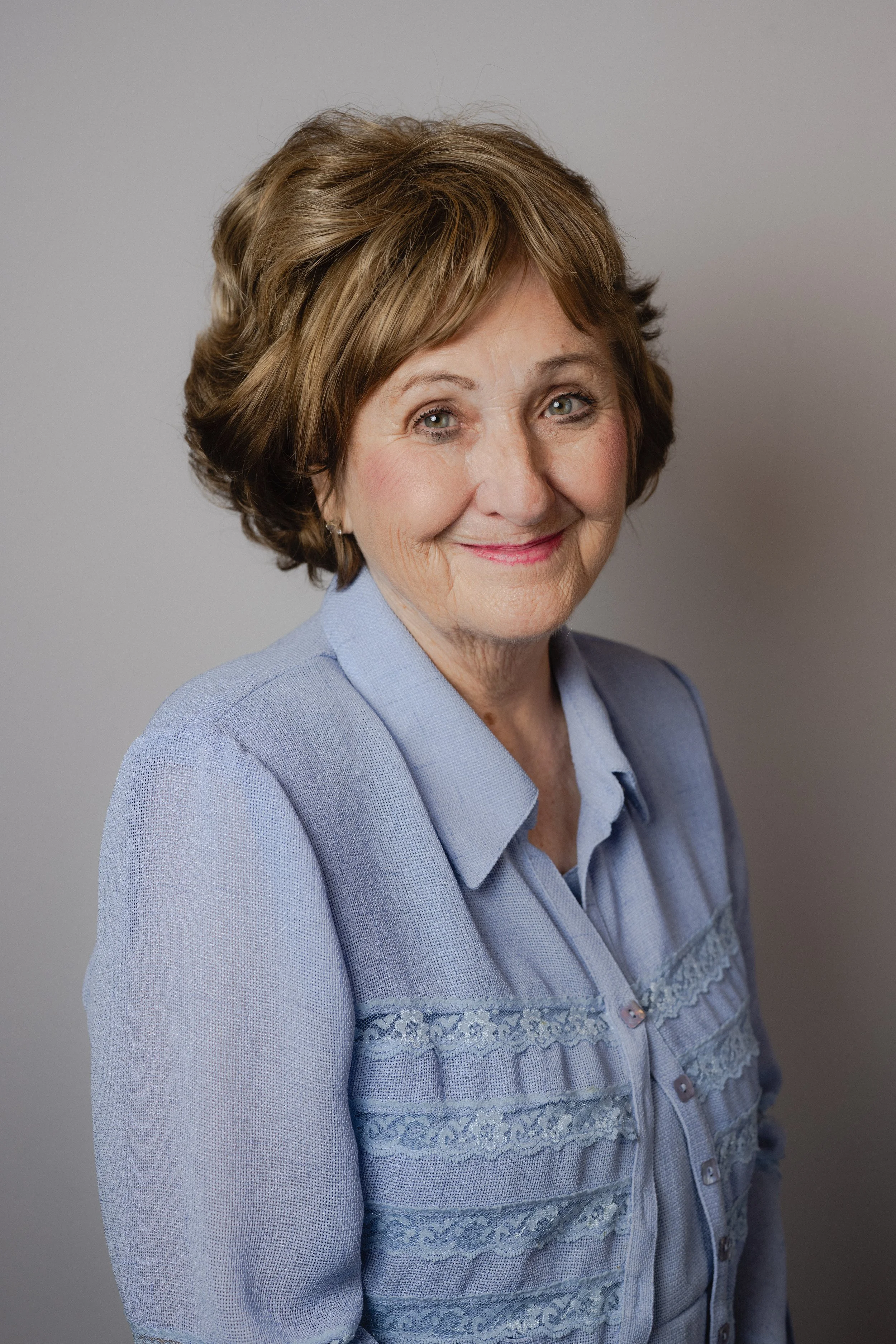 Portrait of an older woman with short, wavy brown hair and grey eyes, wearing a light blue blazer and nice shirt, smiling softly in front of a plain grey background.