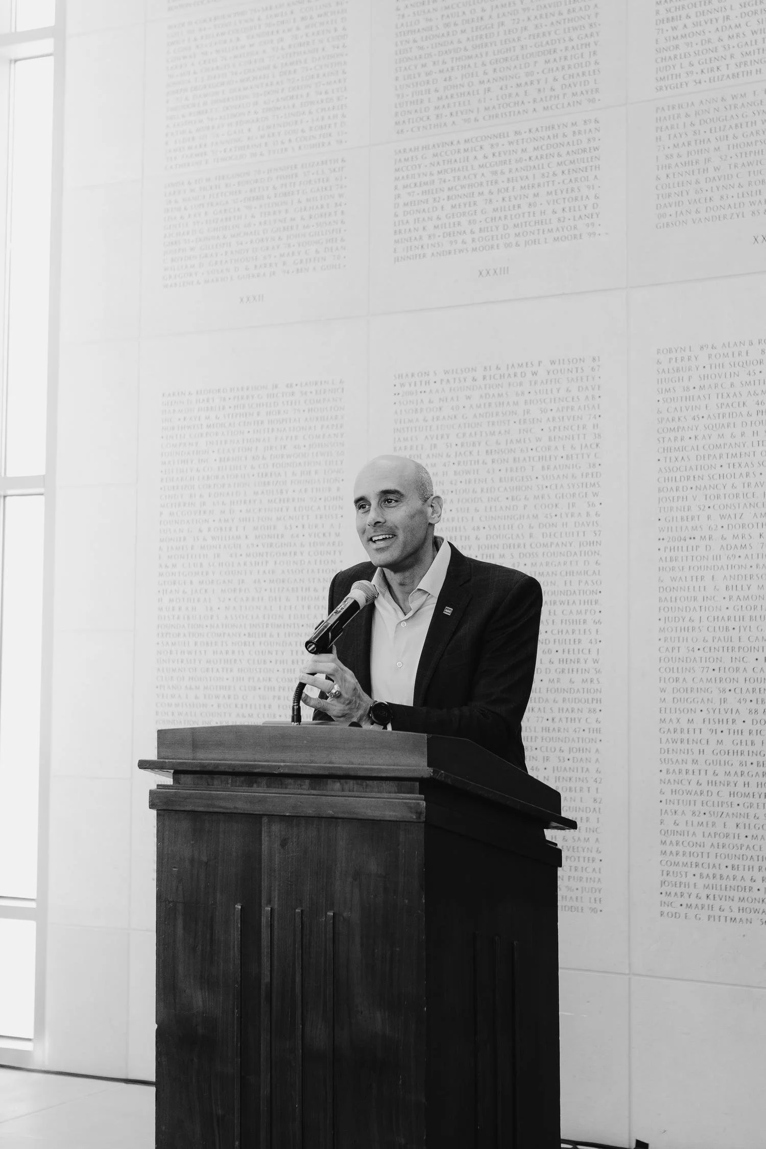 A man in a suit is speaking into a microphone at a podium. Behind him is a wall with lists of names engraved in a large, rectangular plaque.