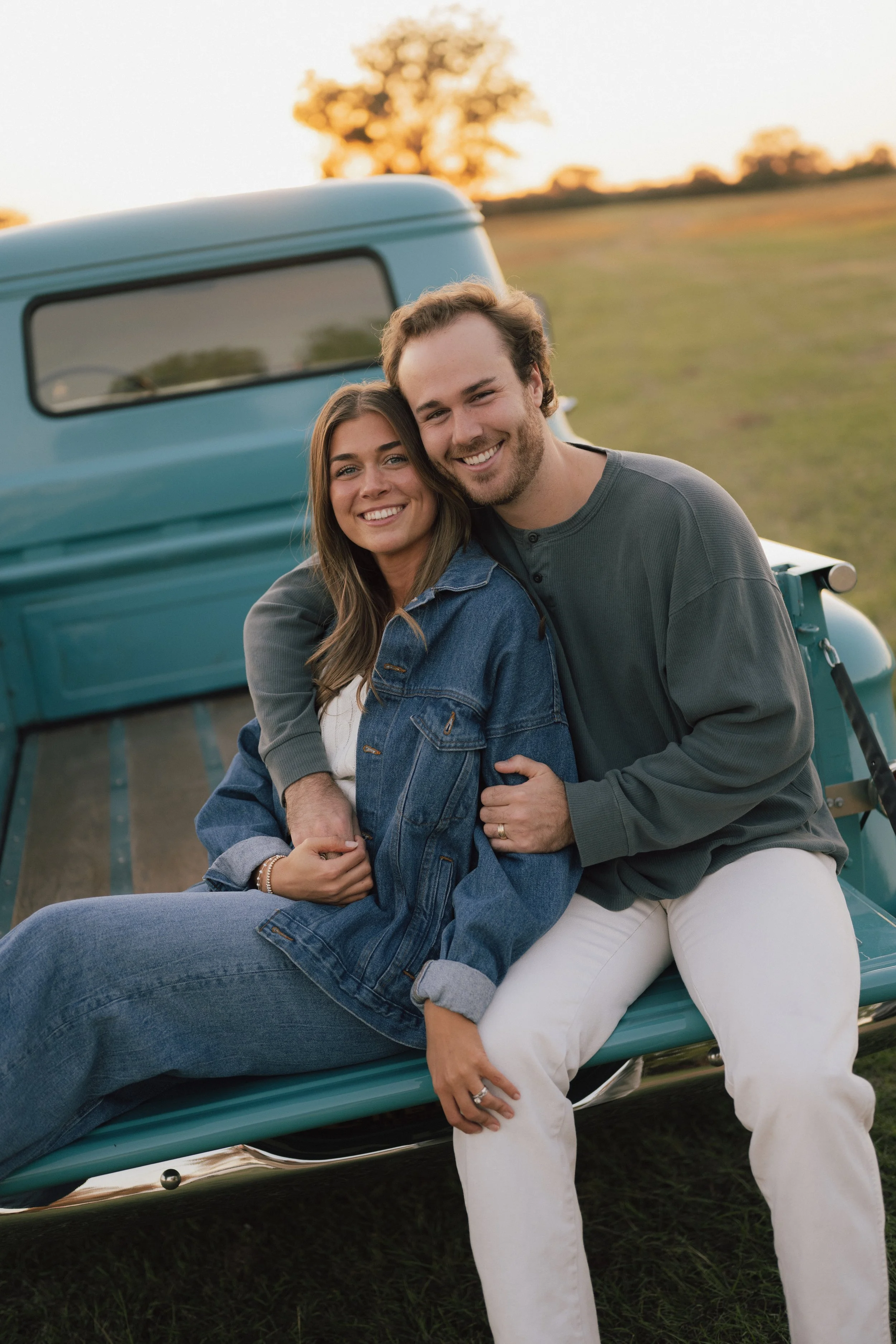 A smiling young woman and man sitting on a vintage blue truck in an open field during sunset, embracing each other.