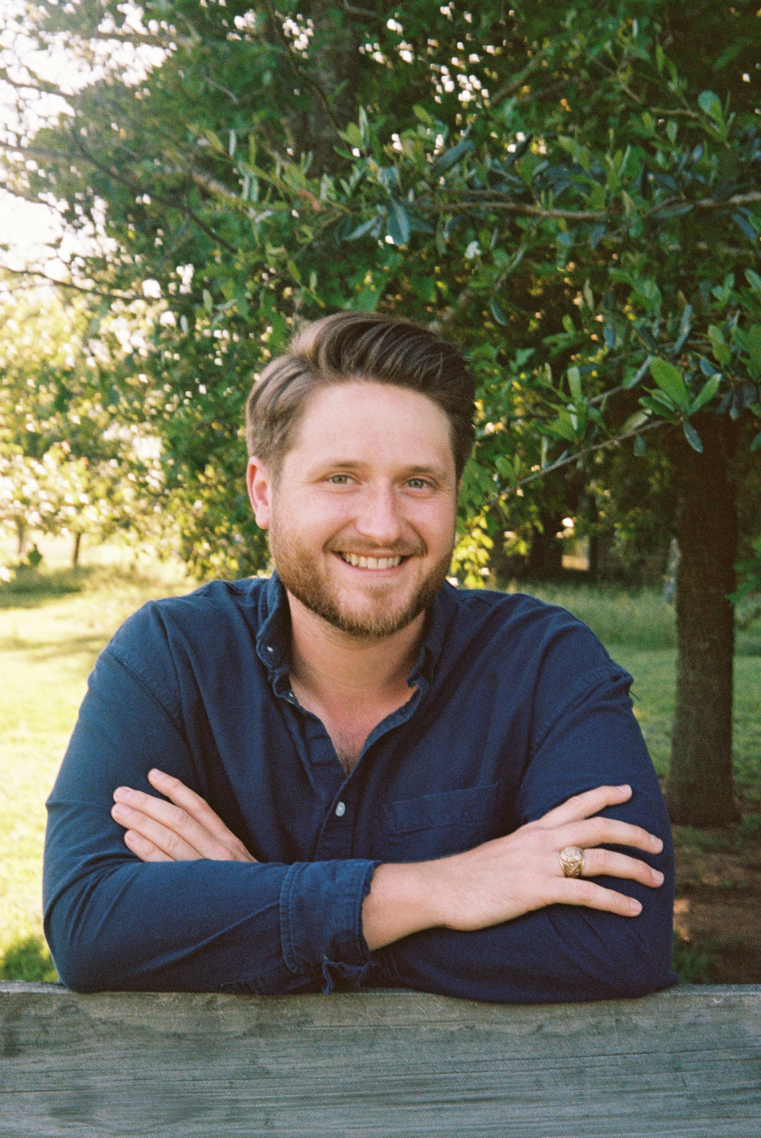 A smiling young man with styled hair and a beard, wearing a dark blue button-up shirt, seated outdoors with arms crossed on a wooden table. Behind him are green trees and shrubbery, illuminated by natural sunlight.